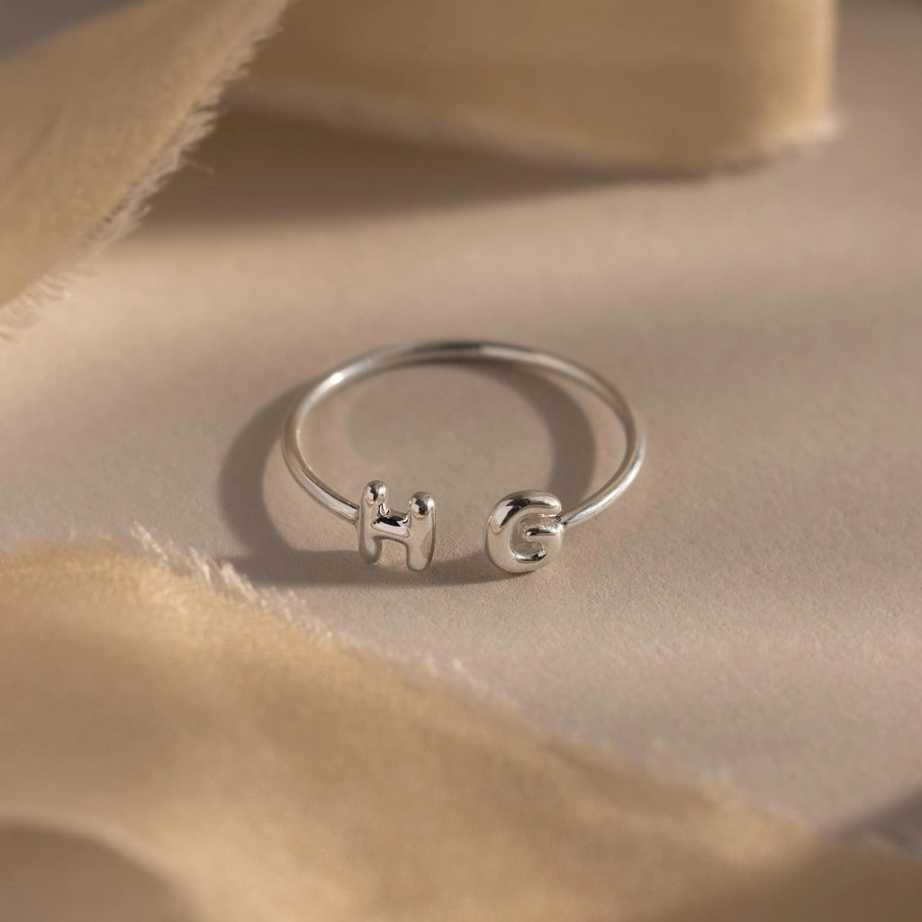 Close-up of a silver personalized ring with bubble letters H and G, styled on neutral beige fabric.