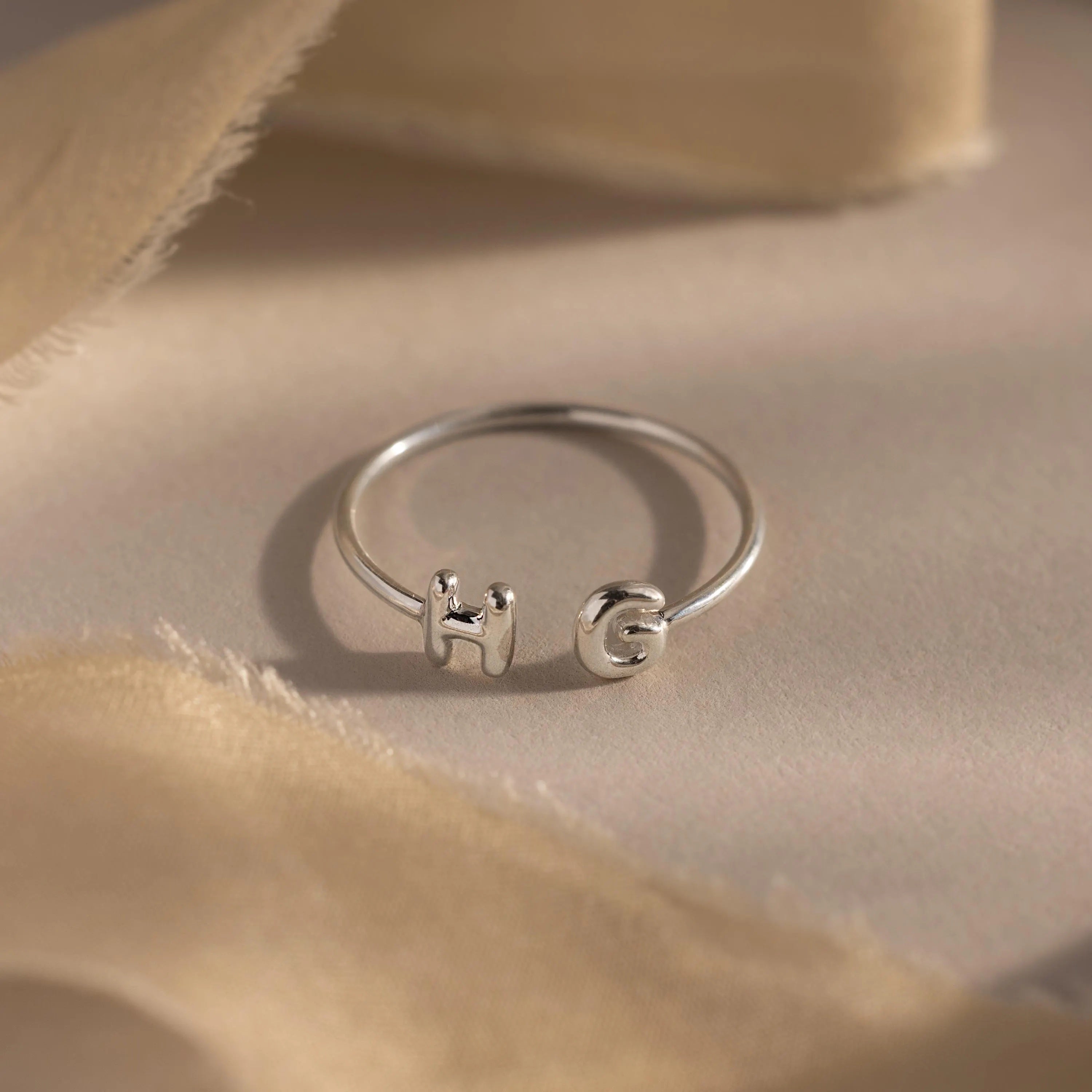 Close-up of a silver personalized ring with bubble letters H and G, styled on neutral beige fabric.