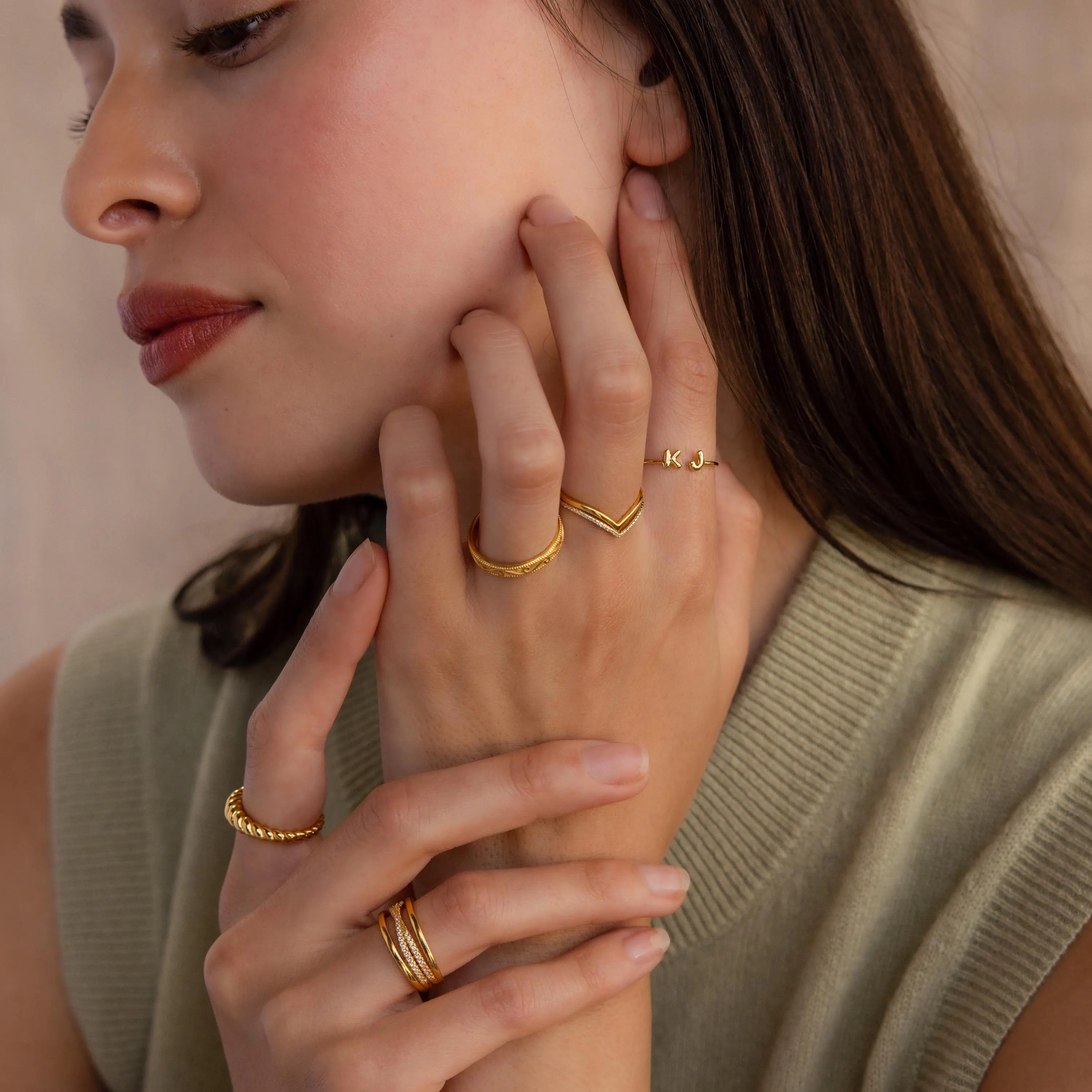 A woman in a sleeveless knit top touches her face while wearing multiple gold rings, including the Duo Bubble Initial Ring in Rose Gold.