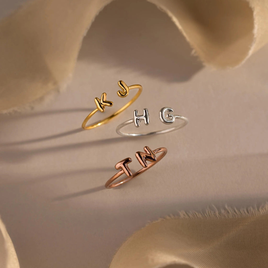 Personalized open rings in three metal finishes—gold, silver, and rose gold—featuring puffy custom initials KJ, HG, and TN, displayed with neutral-toned fabric.