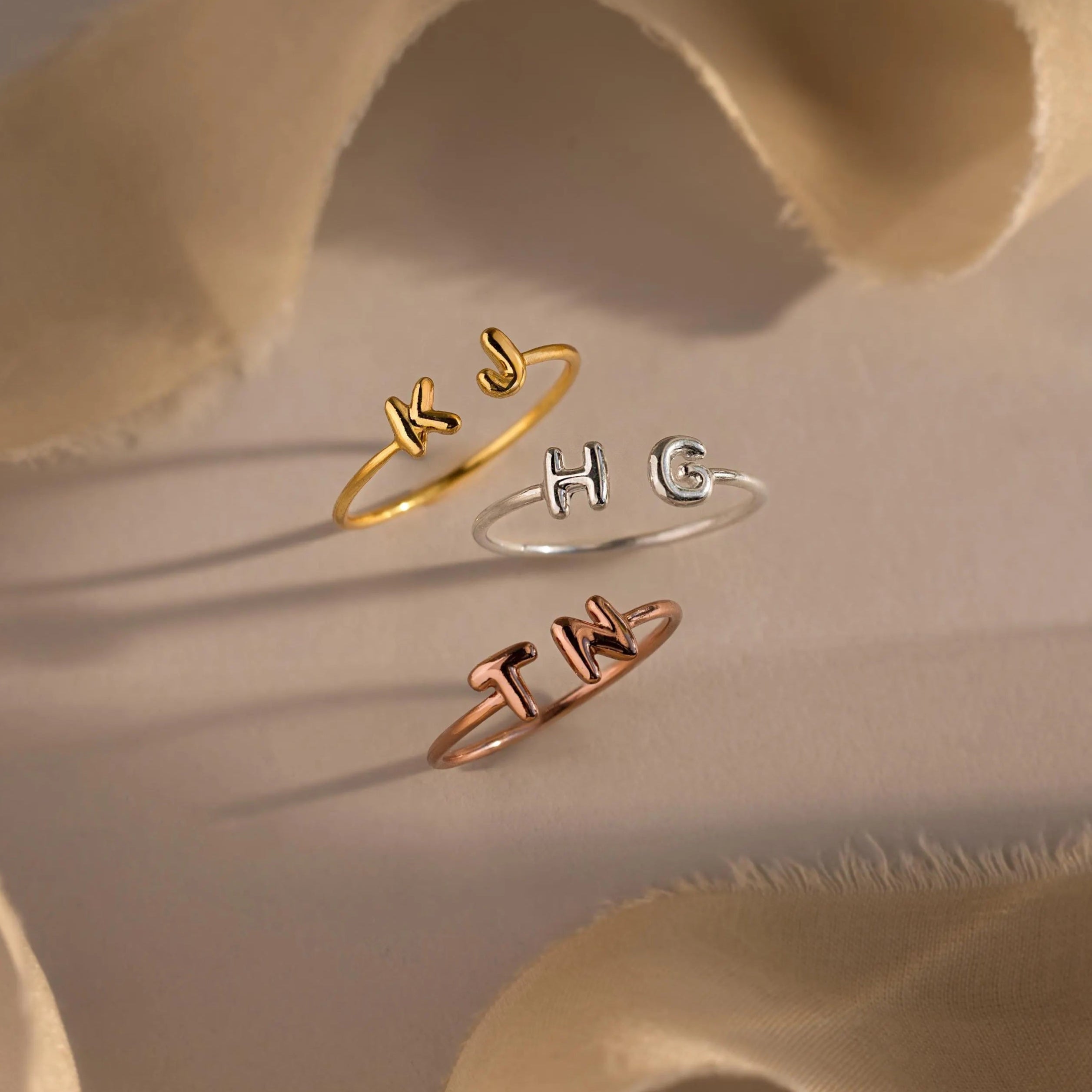 Personalized open rings in three metal finishes—gold, silver, and rose gold—featuring puffy custom initials KJ, HG, and TN, displayed with neutral-toned fabric.
