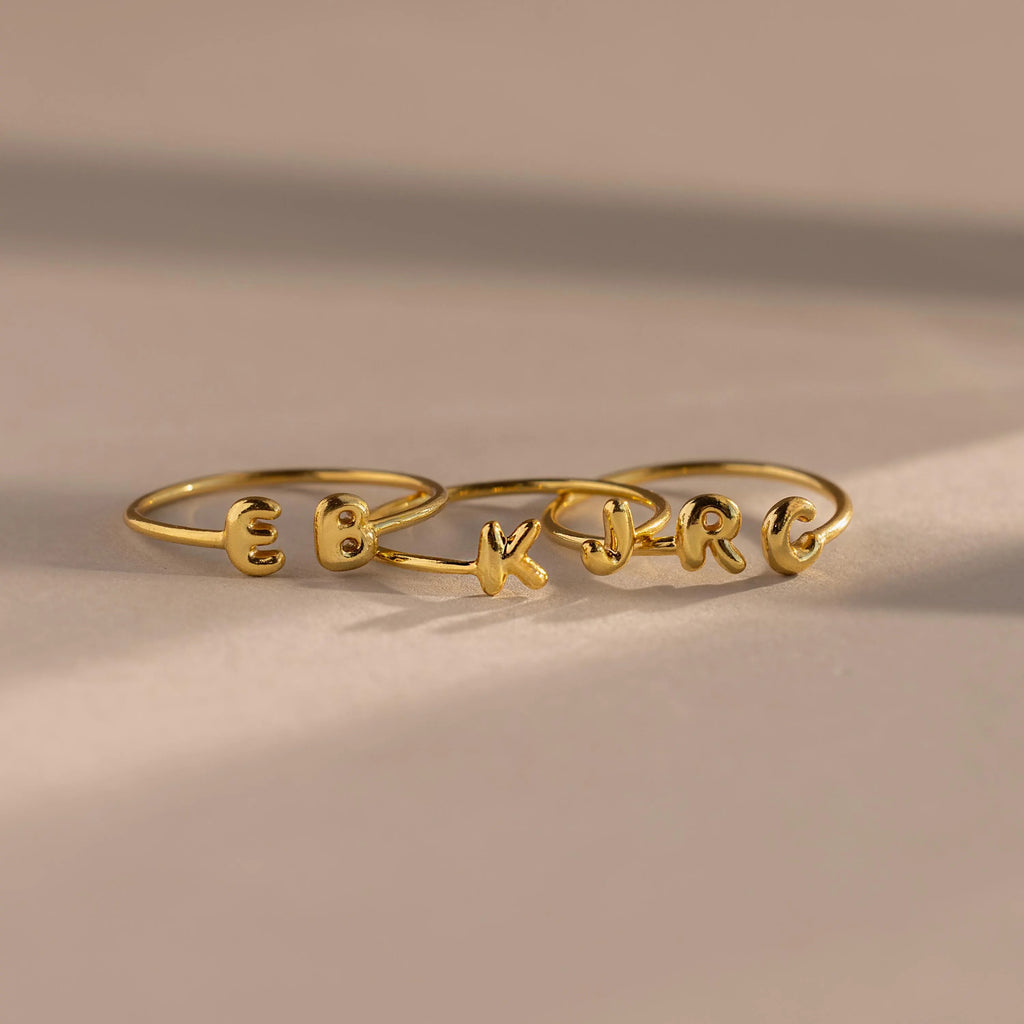 Set of custom gold initial rings with rounded puffy letters, spelling E, B, K, J, R, and C, arranged side by side on a neutral surface.