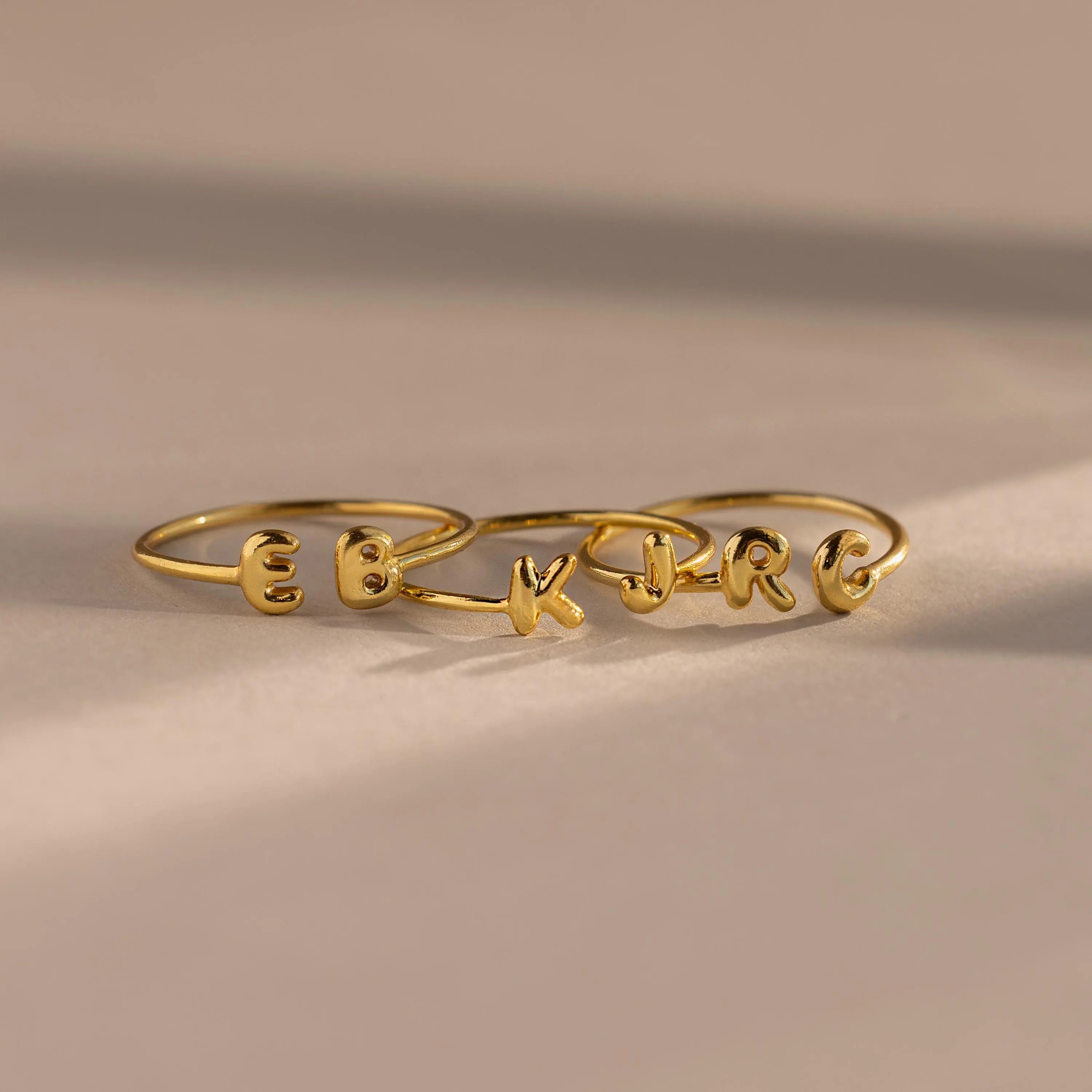 Set of custom gold initial rings with rounded puffy letters, spelling E, B, K, J, R, and C, arranged side by side on a neutral surface.