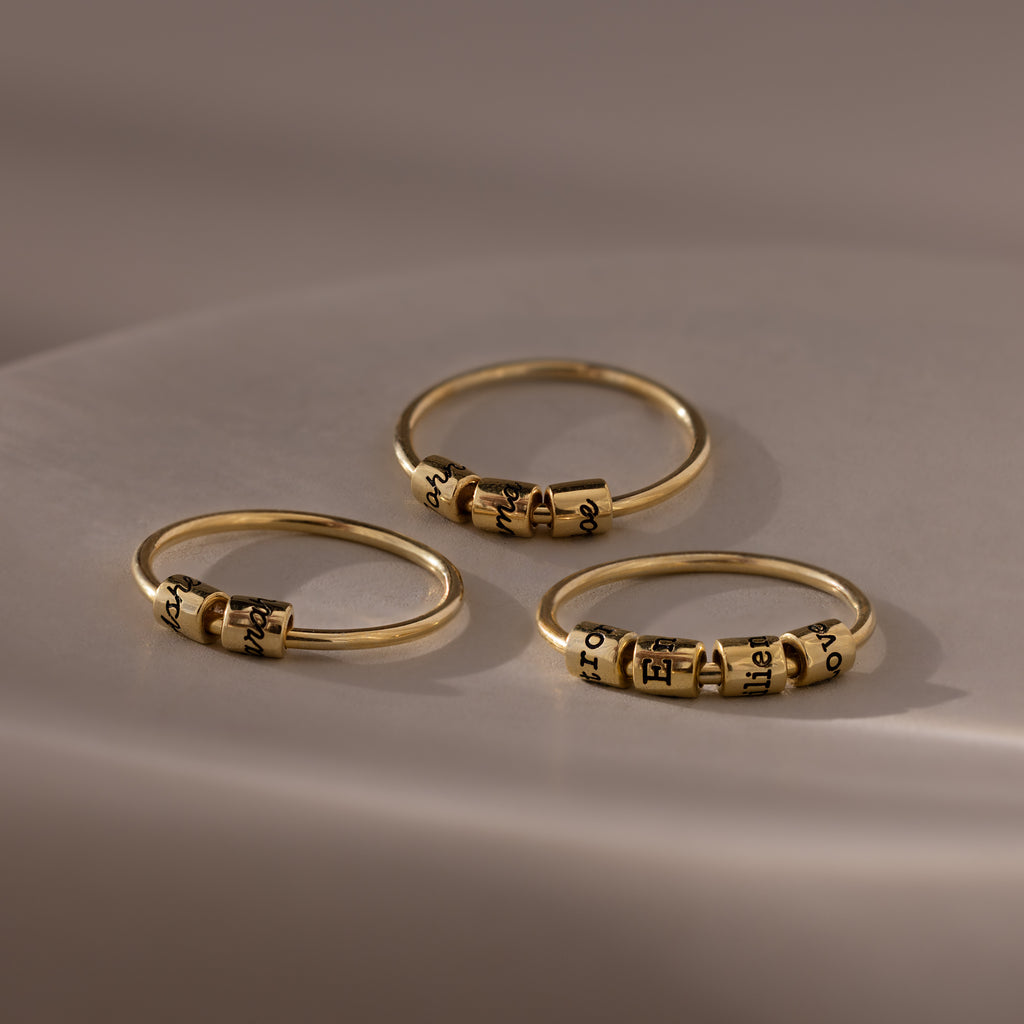 Three Beaded Multiple Name Rings in 18K Gold, each featuring small engraved beads, rest on a smooth, softly lit surface.