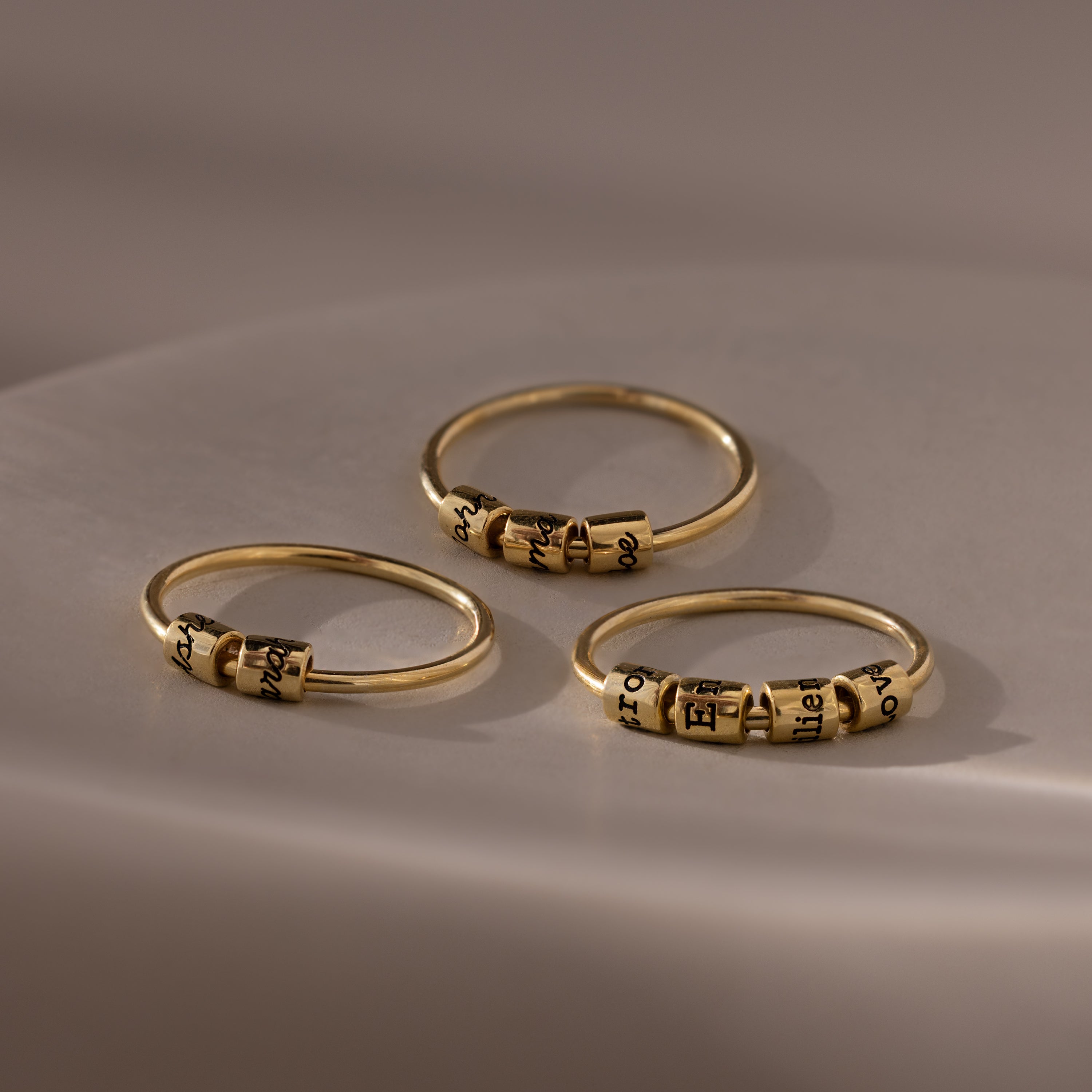 Three Beaded Multiple Name Rings in 18K Gold, each featuring small engraved beads, rest on a smooth, softly lit surface.
