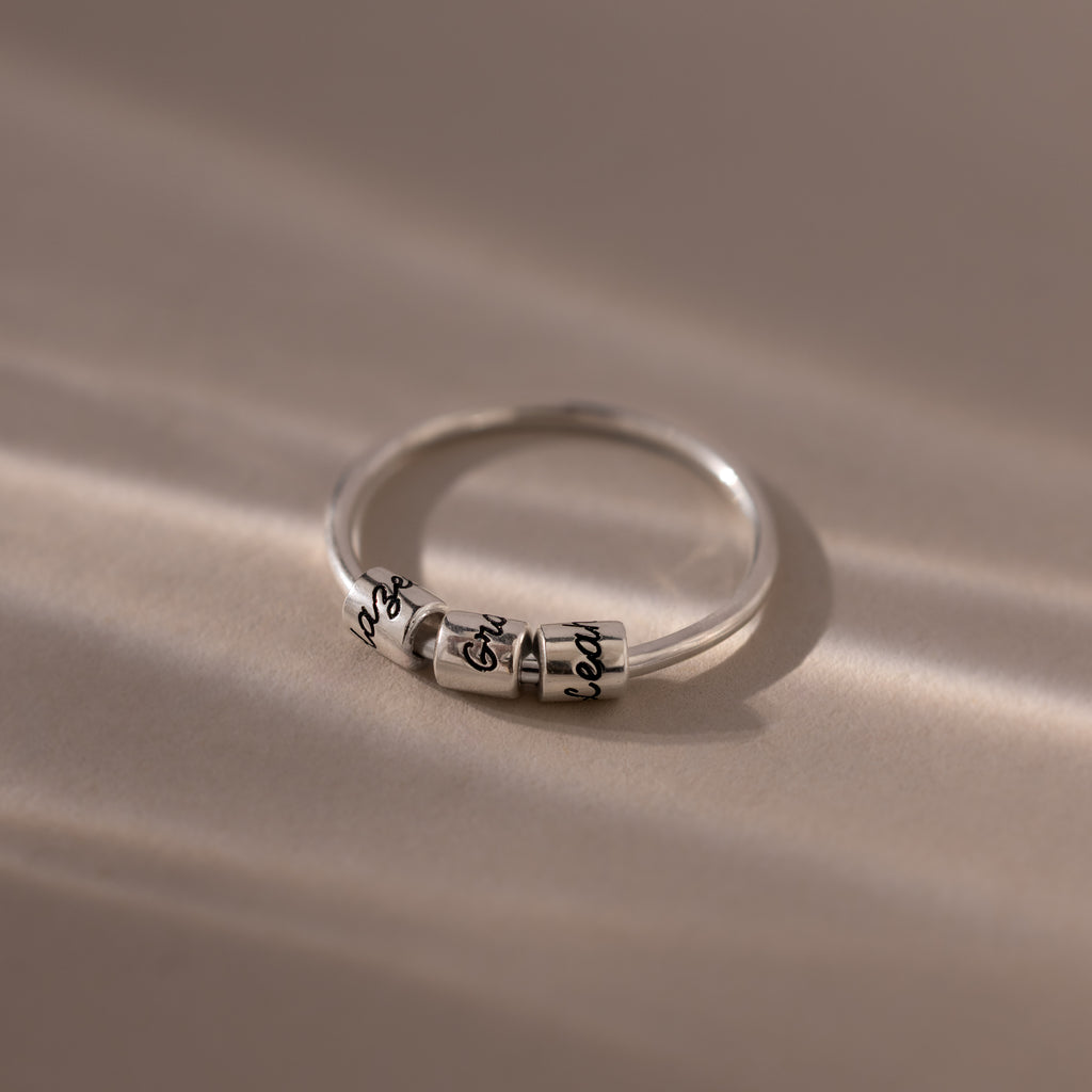 The Beaded Multiple Name Ring in Sterling Silver, featuring delicate engraved beads, rests on a soft beige surface with gentle light and shadows emphasizing its personalized design.