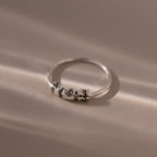 The Beaded Multiple Name Ring in Sterling Silver, featuring delicate engraved beads, rests on a soft beige surface with gentle light and shadows emphasizing its personalized design.