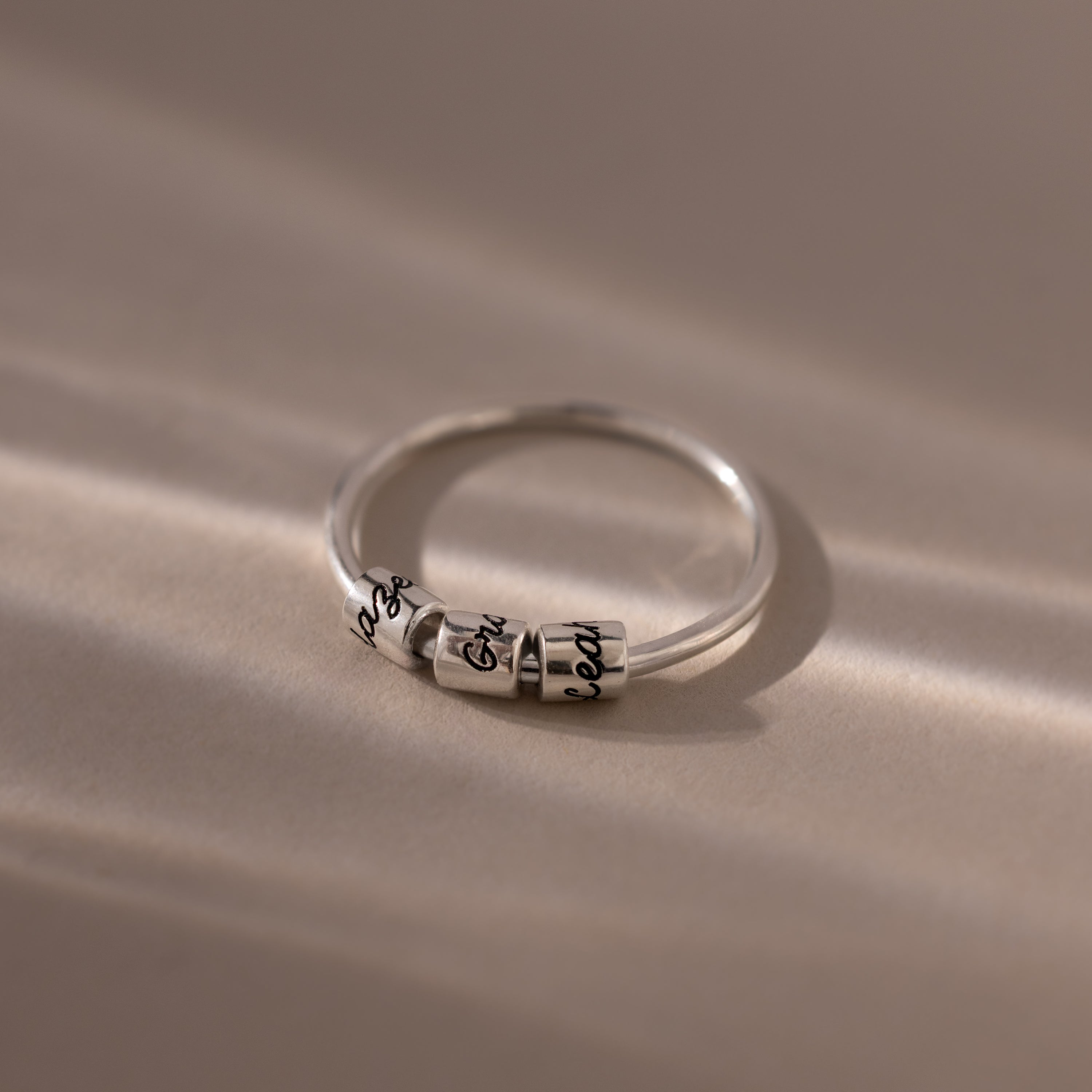 A Beaded Multiple Name Ring, customized with engraved names, sits elegantly on a softly lit, smooth surface.
