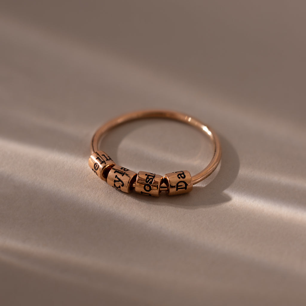 A Beaded Multiple Name Ring in Rose Gold with several engraved spinning letter beads, displayed on a smooth, softly lit surface.