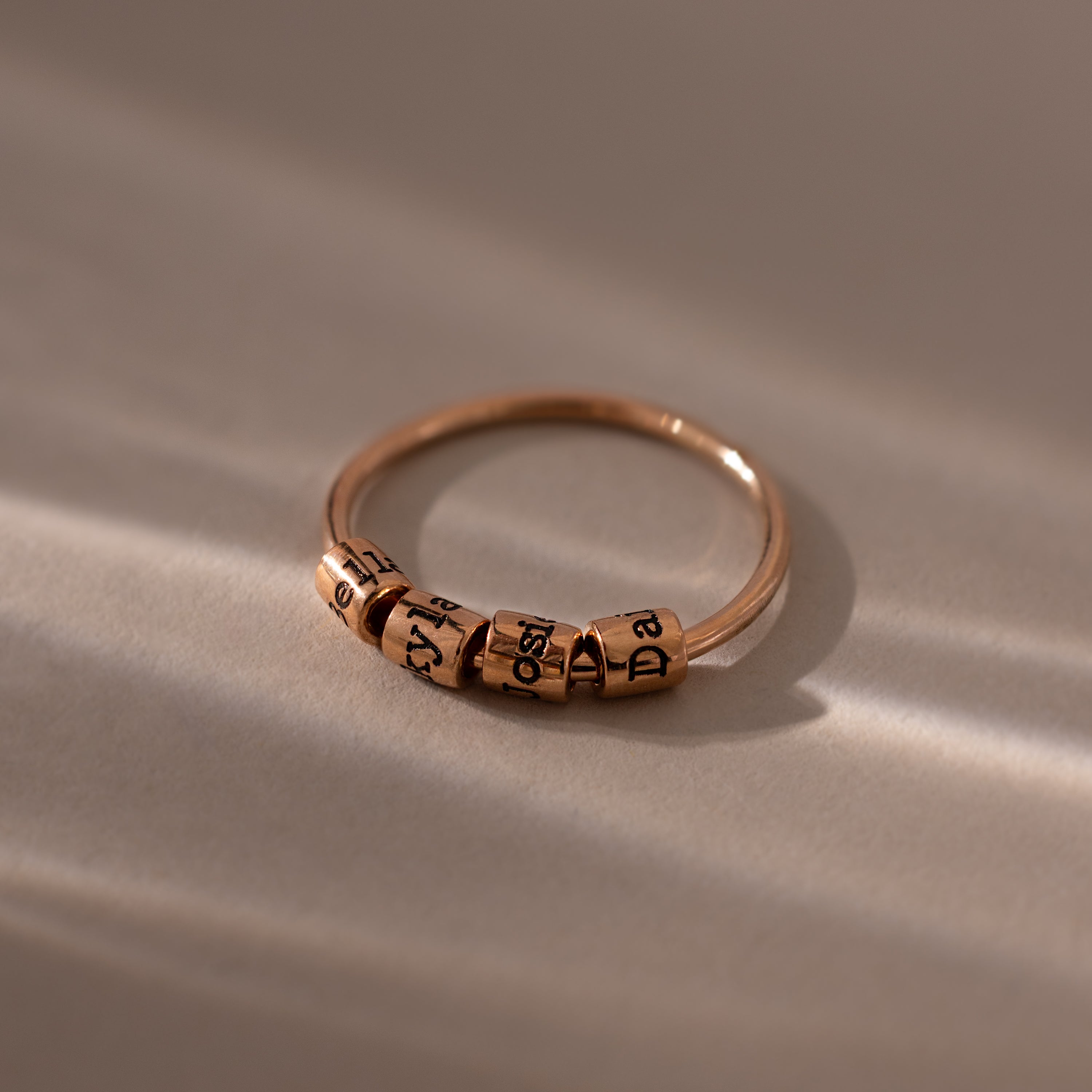 A Beaded Multiple Name Ring in Rose Gold with several engraved spinning letter beads, displayed on a smooth, softly lit surface.