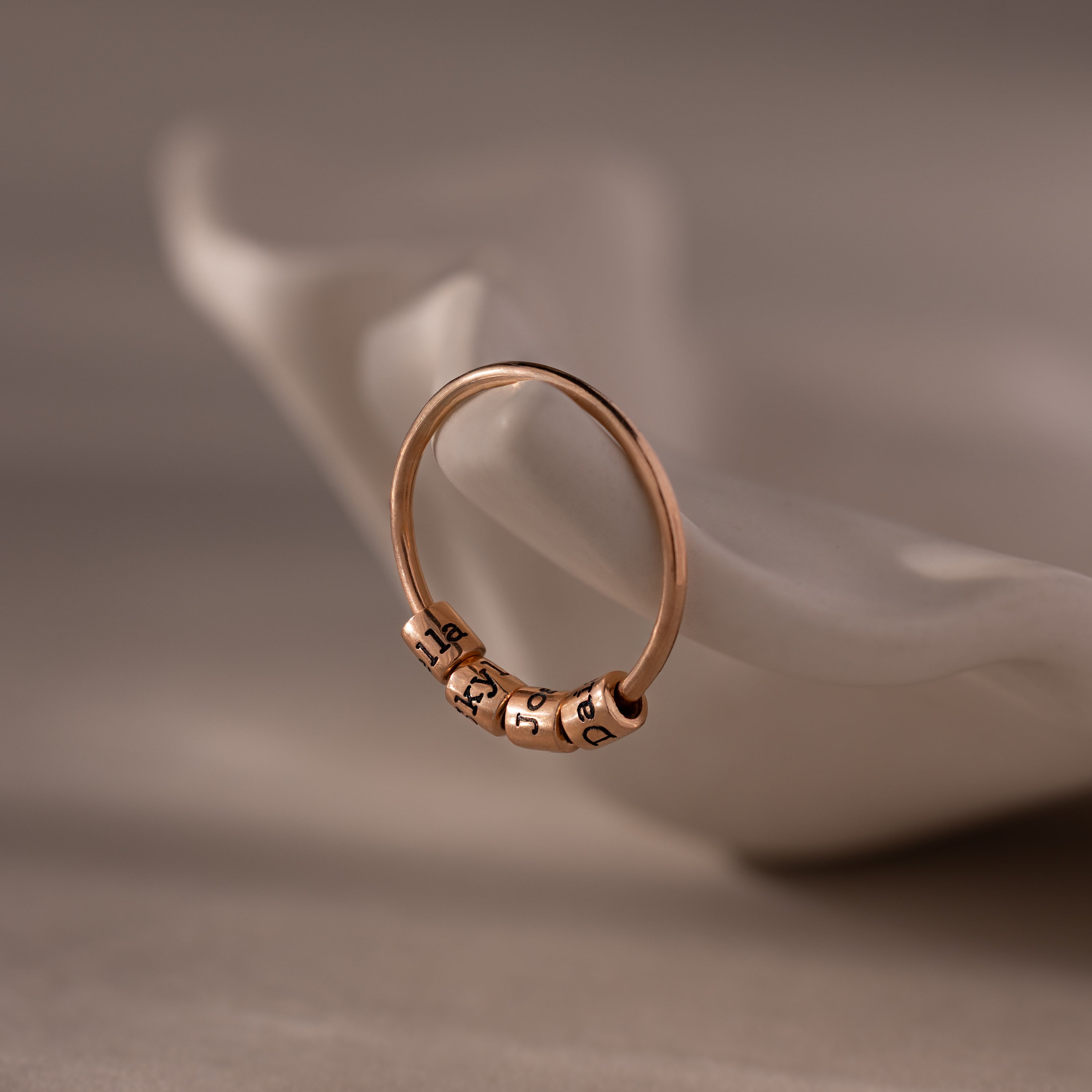 A Beaded Multiple Name Ring with engraved name charms rests on a white, curved ceramic dish.