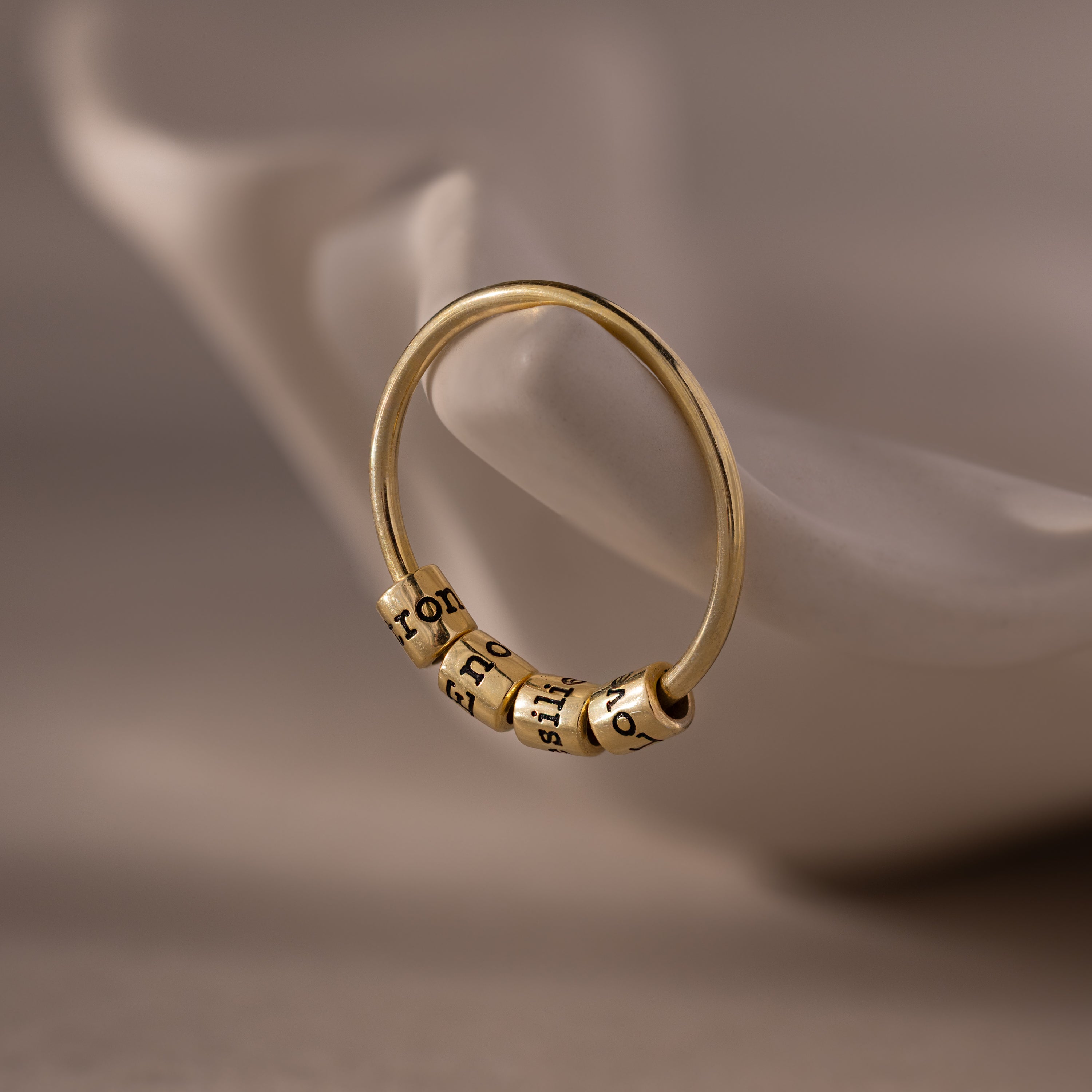 The Beaded Multiple Name Ring, featuring three engraved beads, is displayed on a white, curved ceramic surface.