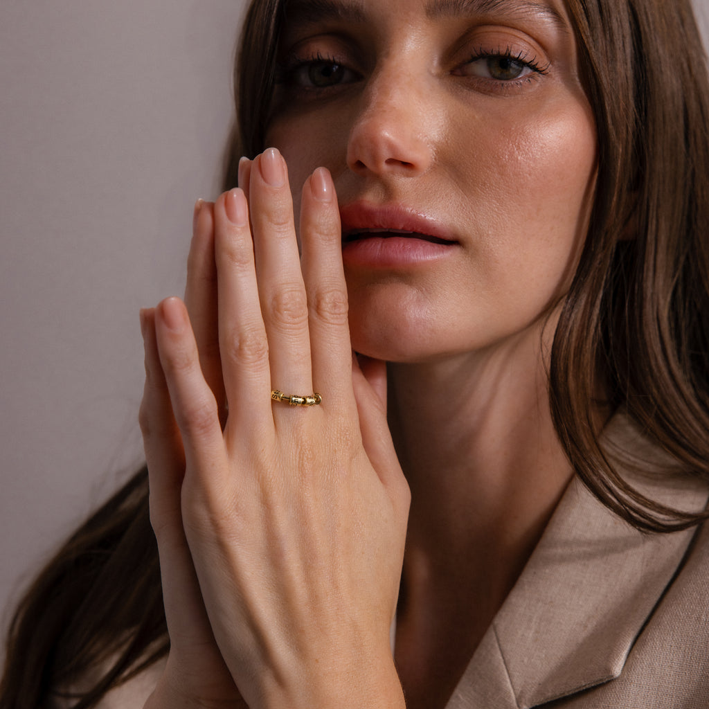 A woman with long brown hair wears a beige blazer and shows off the Beaded Multiple Name Ring in 18K Gold on her hand as she looks at the camera.