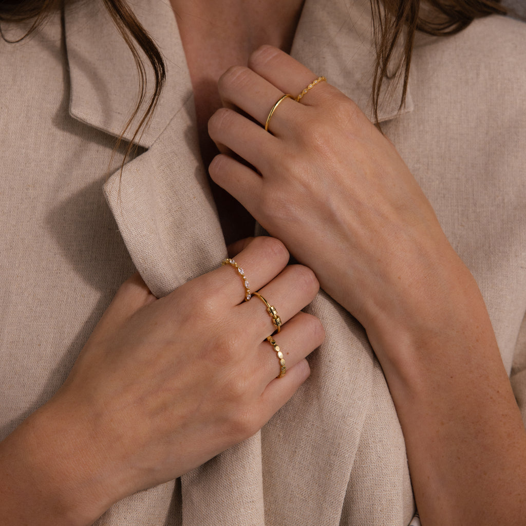 Close-up of hands wearing the Beaded Multiple Name Ring and several gold bands, holding a beige fabric blazer near the collar.