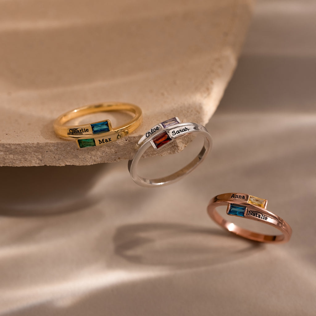 The Toi et Moi Baguette Name Ring in Rose Gold, a personalized name ring with a chic colored gemstone, is elegantly displayed on a beige stone surface.