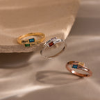 The Toi et Moi Baguette Name Ring in Rose Gold, a personalized name ring with a chic colored gemstone, is elegantly displayed on a beige stone surface.