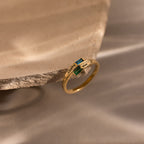 The Toi et Moi Baguette Name Ring in 18K Gold, featuring two gemstones and engraved names, is displayed on the edge of a textured stone surface.