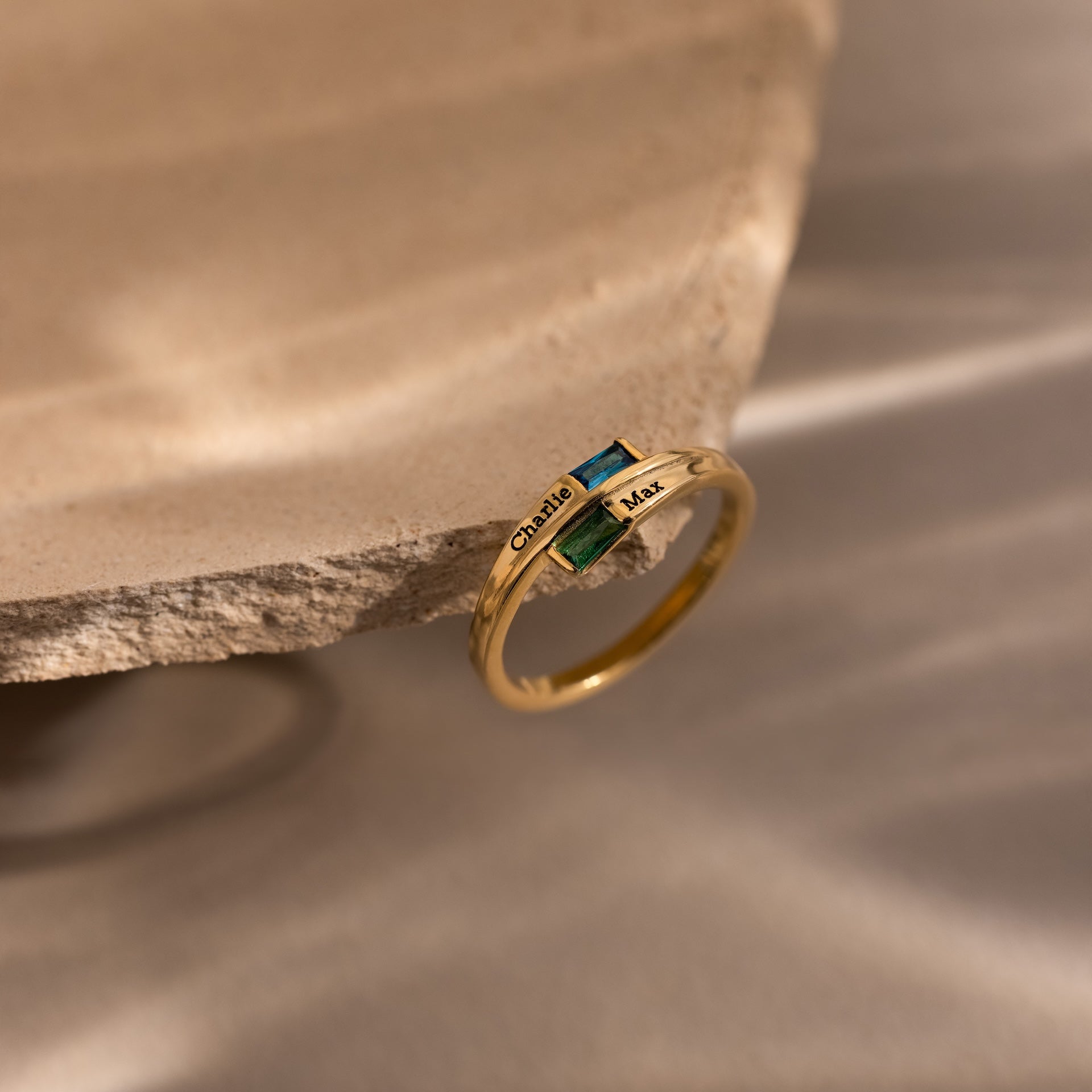 The Toi et Moi Baguette Name Ring in 18K Gold, featuring two gemstones and engraved names, is displayed on the edge of a textured stone surface.