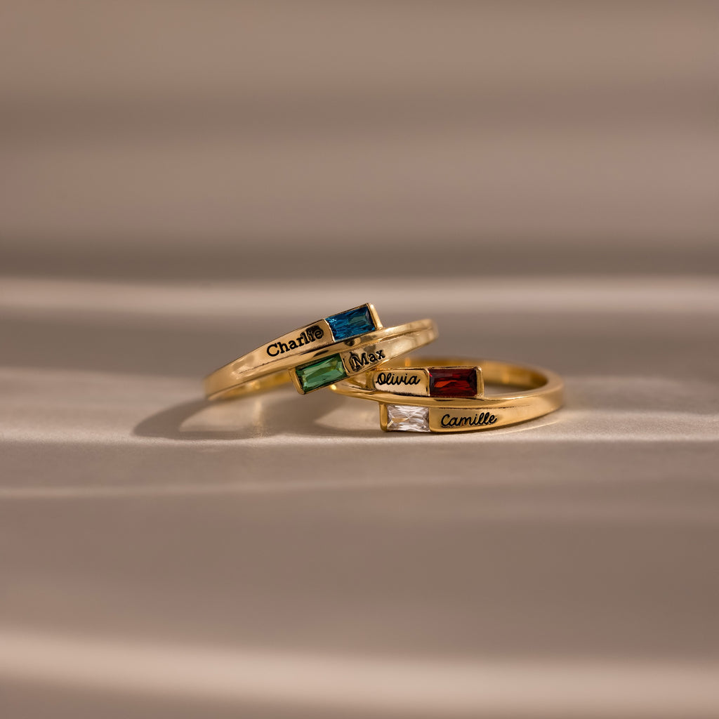 The Toi et Moi Baguette Name Ring, along with two other gold rings adorned with colorful gemstones and engraved names, rests elegantly on a soft beige surface.