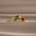 The Toi et Moi Baguette Name Ring, along with two other gold rings adorned with colorful gemstones and engraved names, rests elegantly on a soft beige surface.