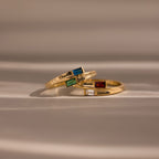 The Toi et Moi Baguette Name Ring, along with two other gold rings adorned with colorful gemstones and engraved names, rests elegantly on a soft beige surface.