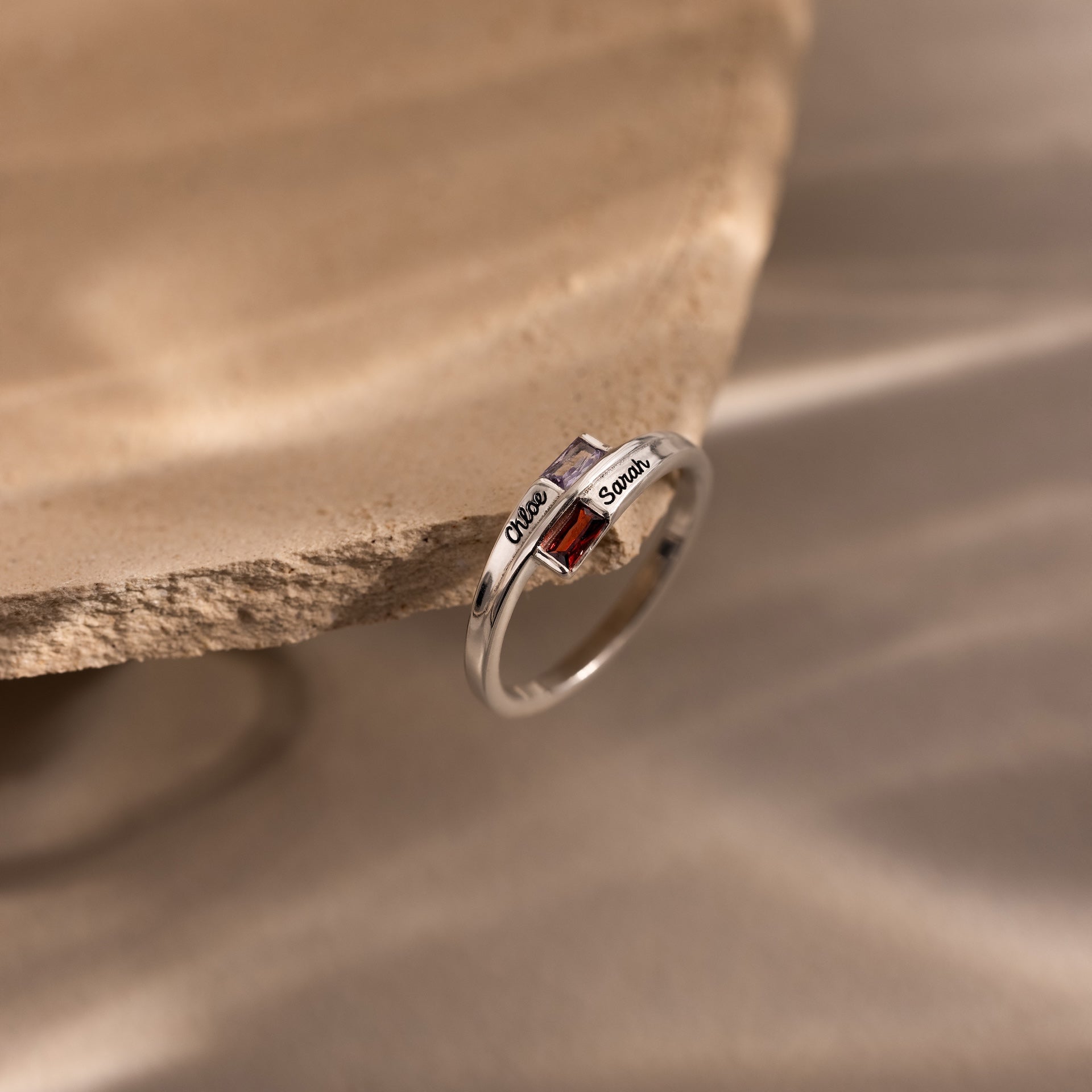 The Toi et Moi Baguette Name Ring in Sterling Silver features two square gemstones and custom engraved names, displayed elegantly on the edge of a stone surface.
