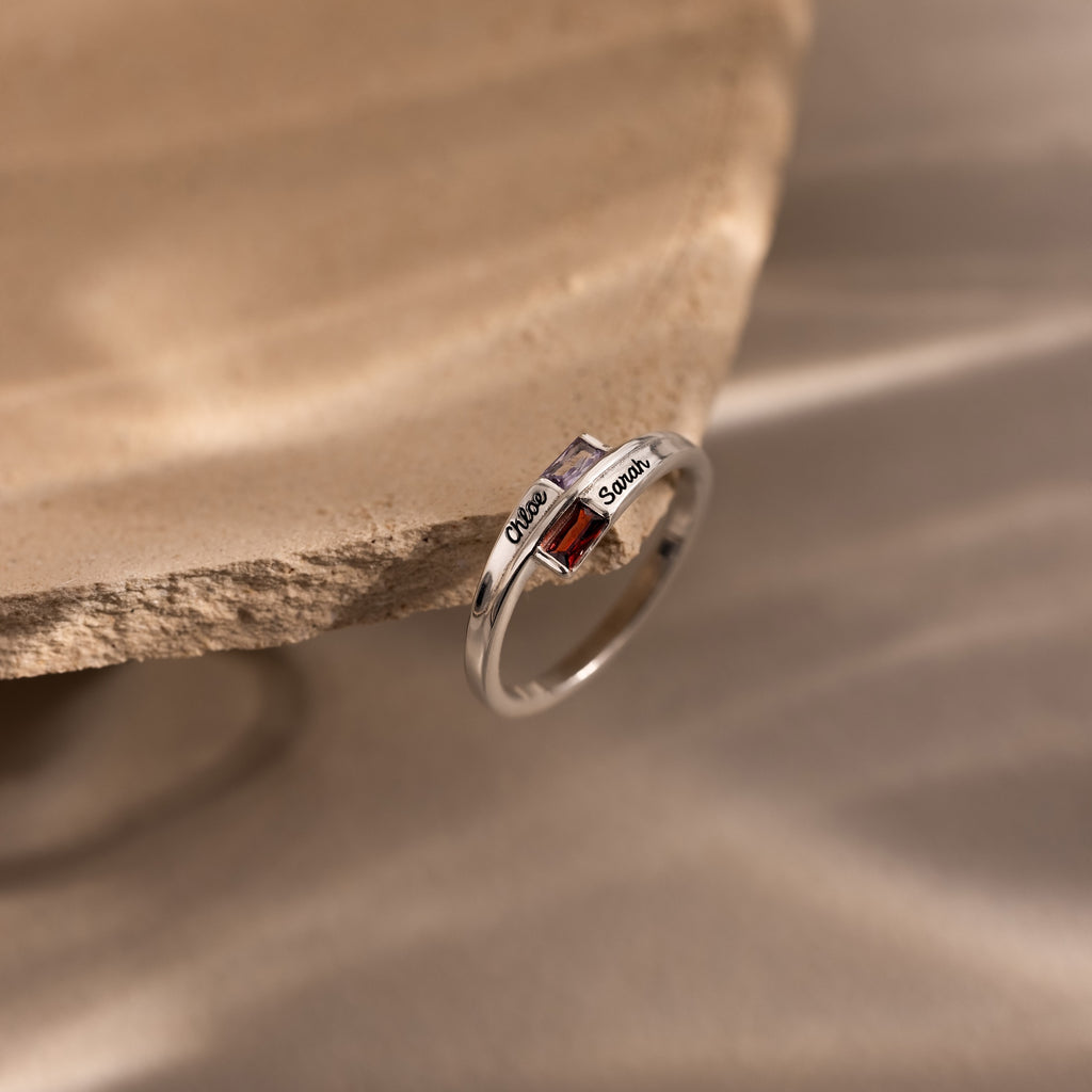The Toi et Moi Baguette Name Ring features two square gemstones with engraved names, inspired by the classic Toi et Moi style—a personalized birthstone ring resting on a textured beige surface.