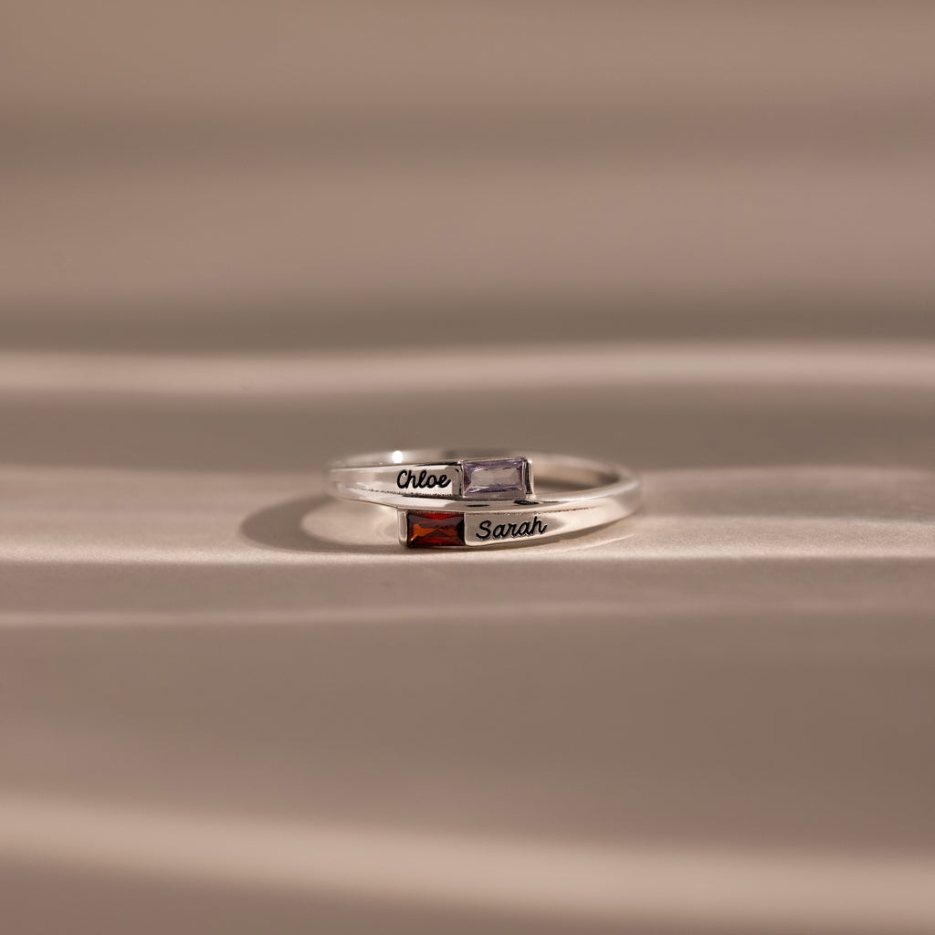 Two Toi et Moi Baguette Name Rings in Sterling Silver, each with colored gemstones and engraved names, Chloe and Sarah, rest on a soft beige surface—a perfect personalized gift for two.