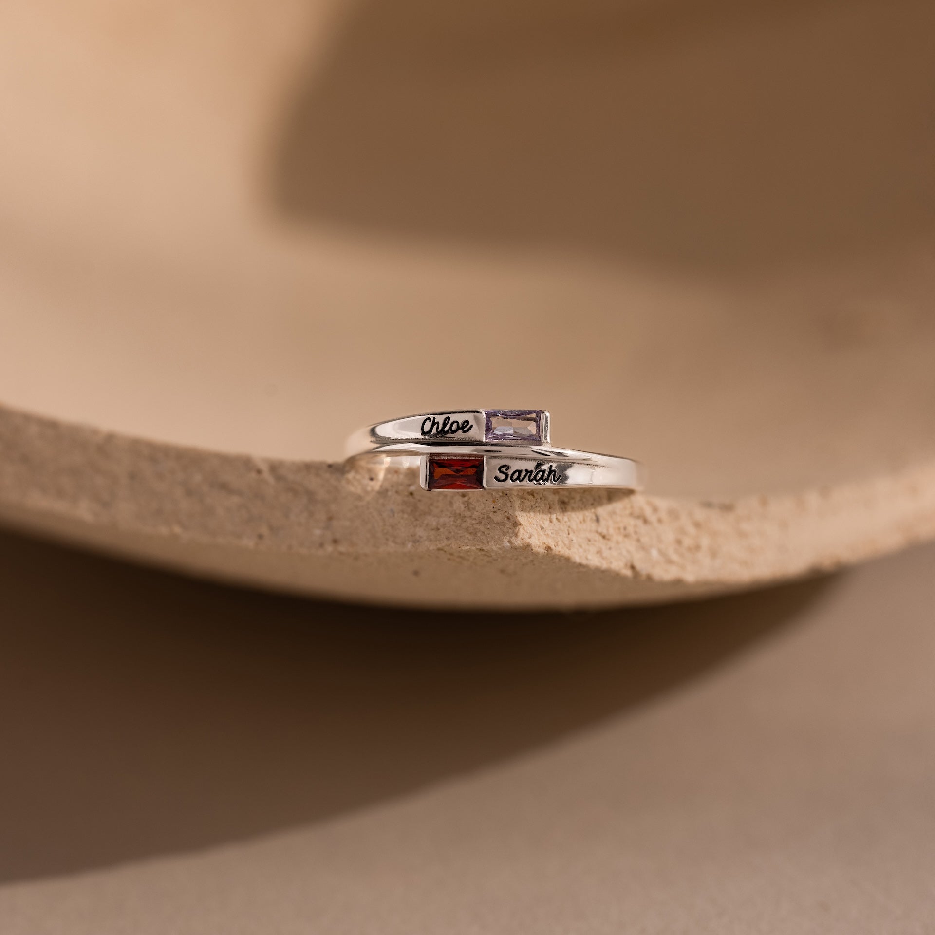 Two Toi et Moi Baguette Name Rings in Sterling Silver, personalized with engraved names Chloe and Sarah plus colorful stones, rest on a beige surface—a unique customizable name ring for any occasion.