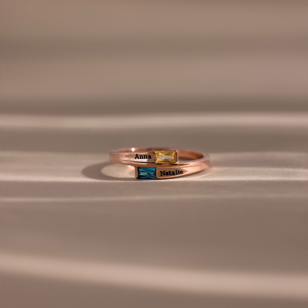 Two Toi et Moi Baguette Name Rings in rose gold, personalized with "Anna" and "Natalie" and accented with colored gemstones, rest on a soft surface—perfect for symbolizing a unique connection.