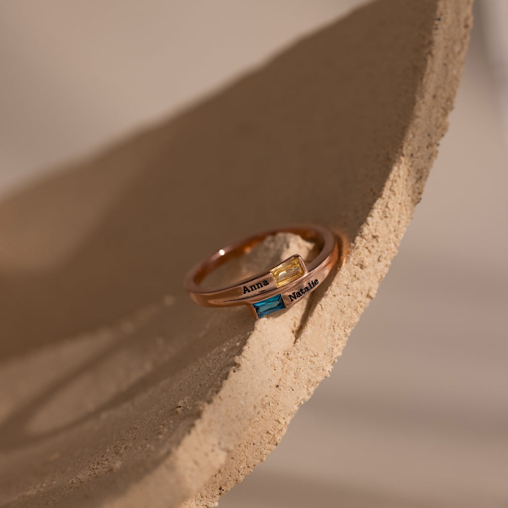 The Toi et Moi Baguette Name Ring in Rose Gold features two colored stones and engraved names Anna and Natalie, elegantly displayed on a textured beige surface.