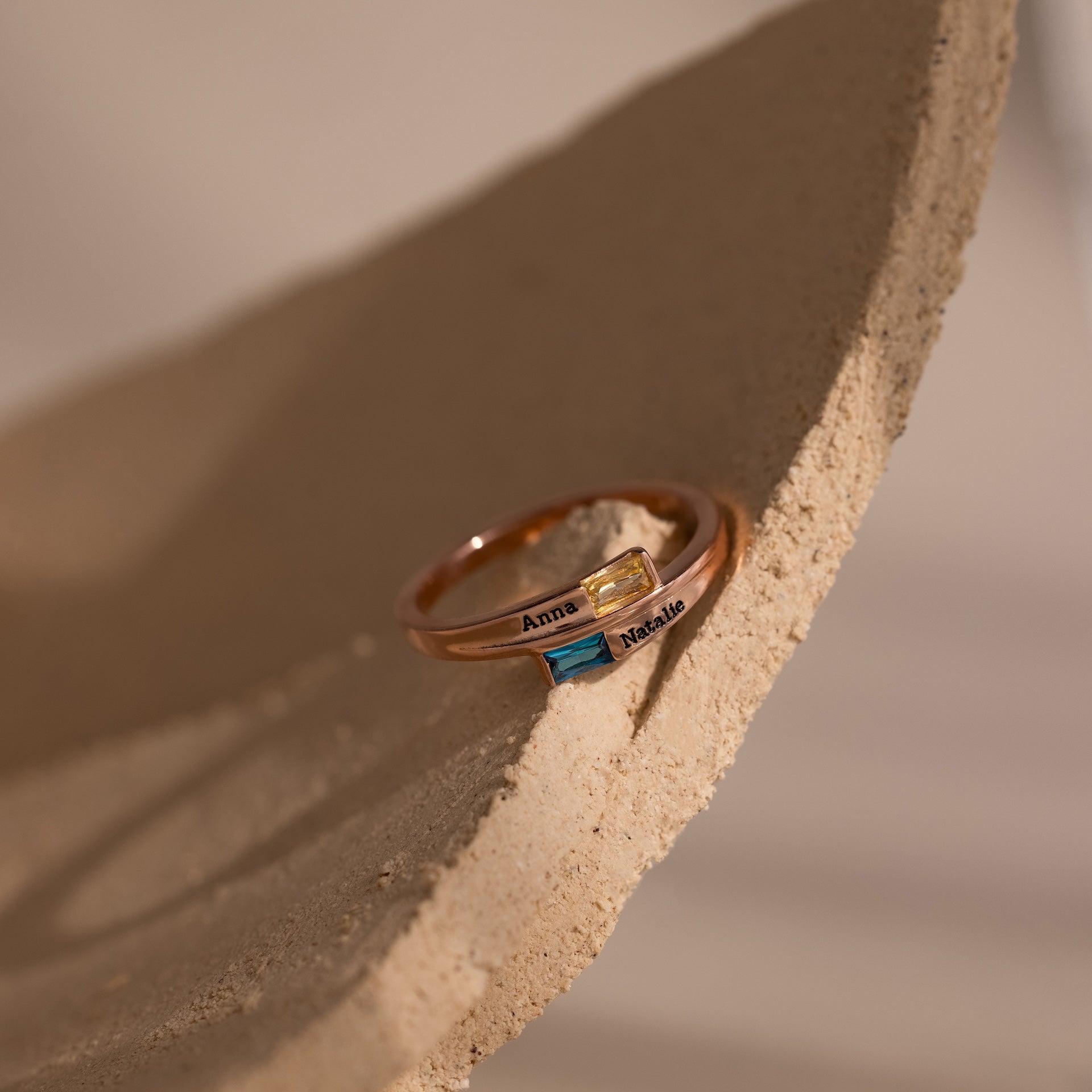 The Toi et Moi Baguette Name Ring in Rose Gold features two colored stones and engraved names Anna and Natalie, elegantly displayed on a textured beige surface.