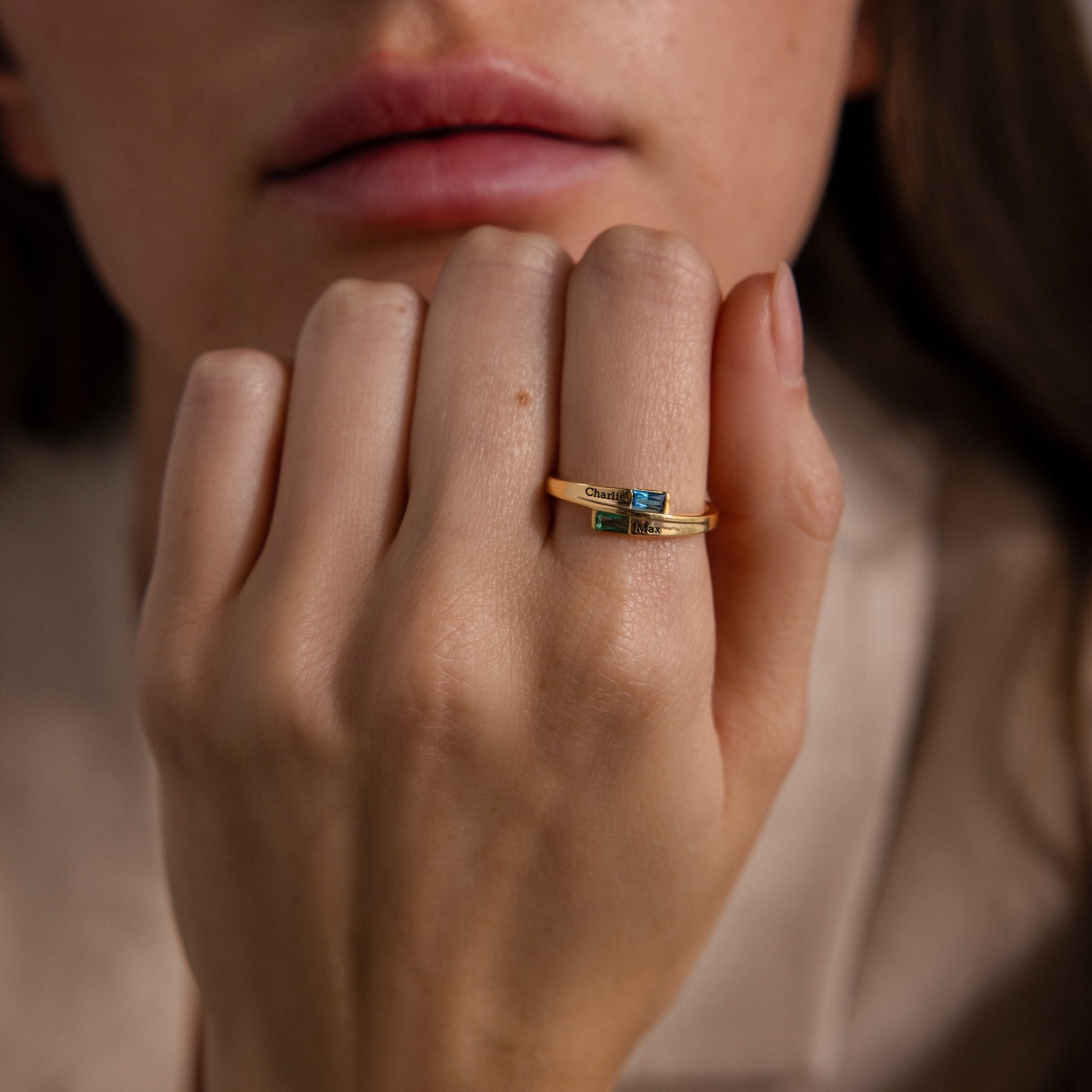 A close-up of a hand with two gold rings, including the Toi et Moi Baguette Name Ring and another featuring a small blue gemstone, held near the lips.