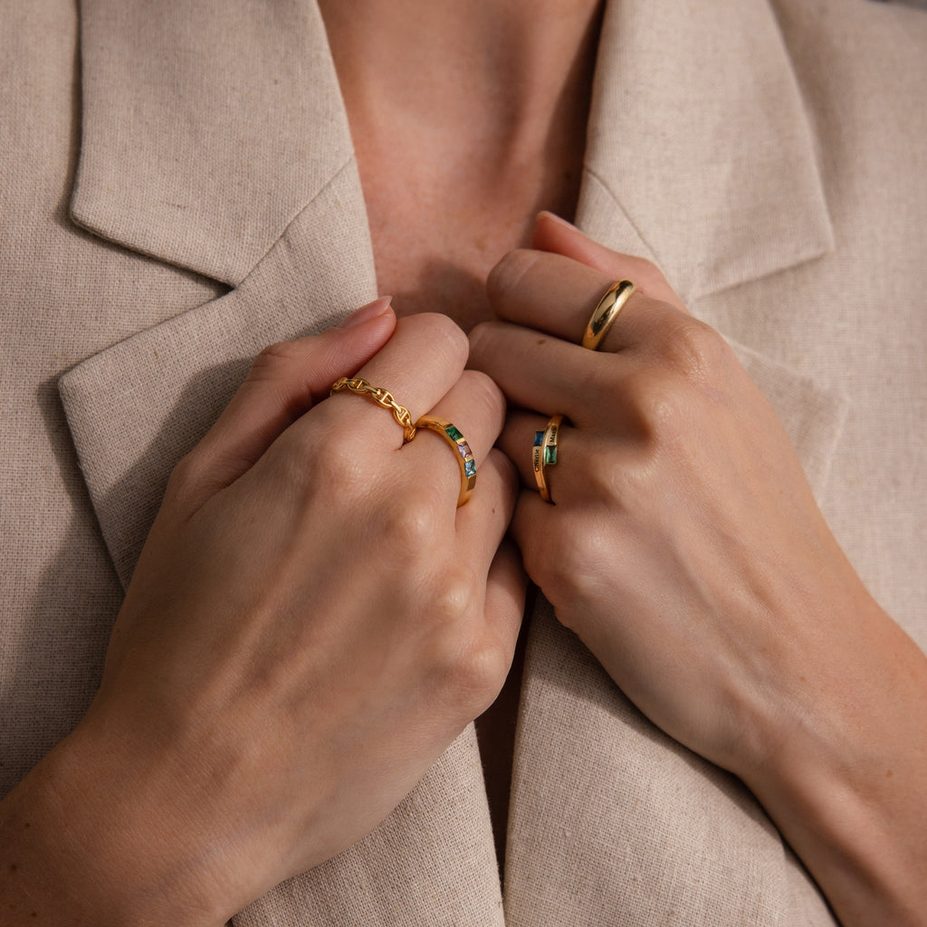 Hands adorned with gold rings, including the Toi et Moi Baguette Name Ring, held near the chest and styled with a beige blazer.