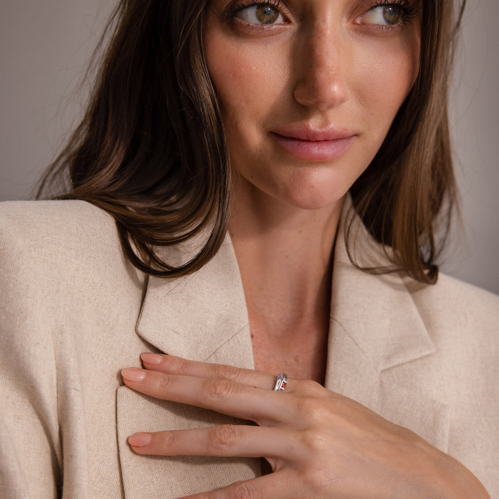 A woman in a beige blazer touches her chest, wearing the Toi et Moi Baguette Name Ring in Sterling Silver, and gazes slightly to the side.