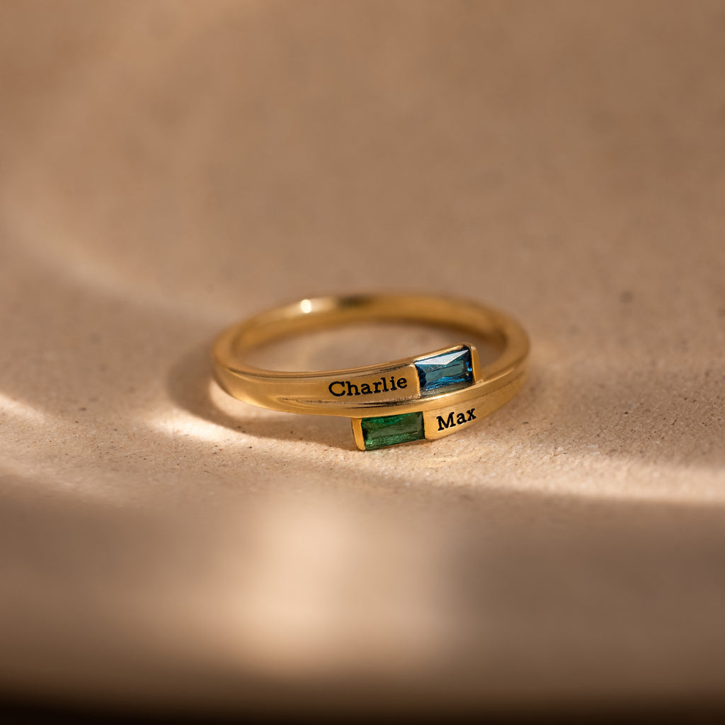 The Toi et Moi Baguette Name Ring in 18K Gold features two rectangular gemstones and engraved names, Charlie and Max, displayed on a beige surface.