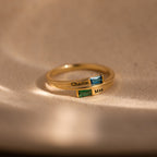 The Toi et Moi Baguette Name Ring in 18K Gold features two rectangular gemstones and engraved names, Charlie and Max, displayed on a beige surface.