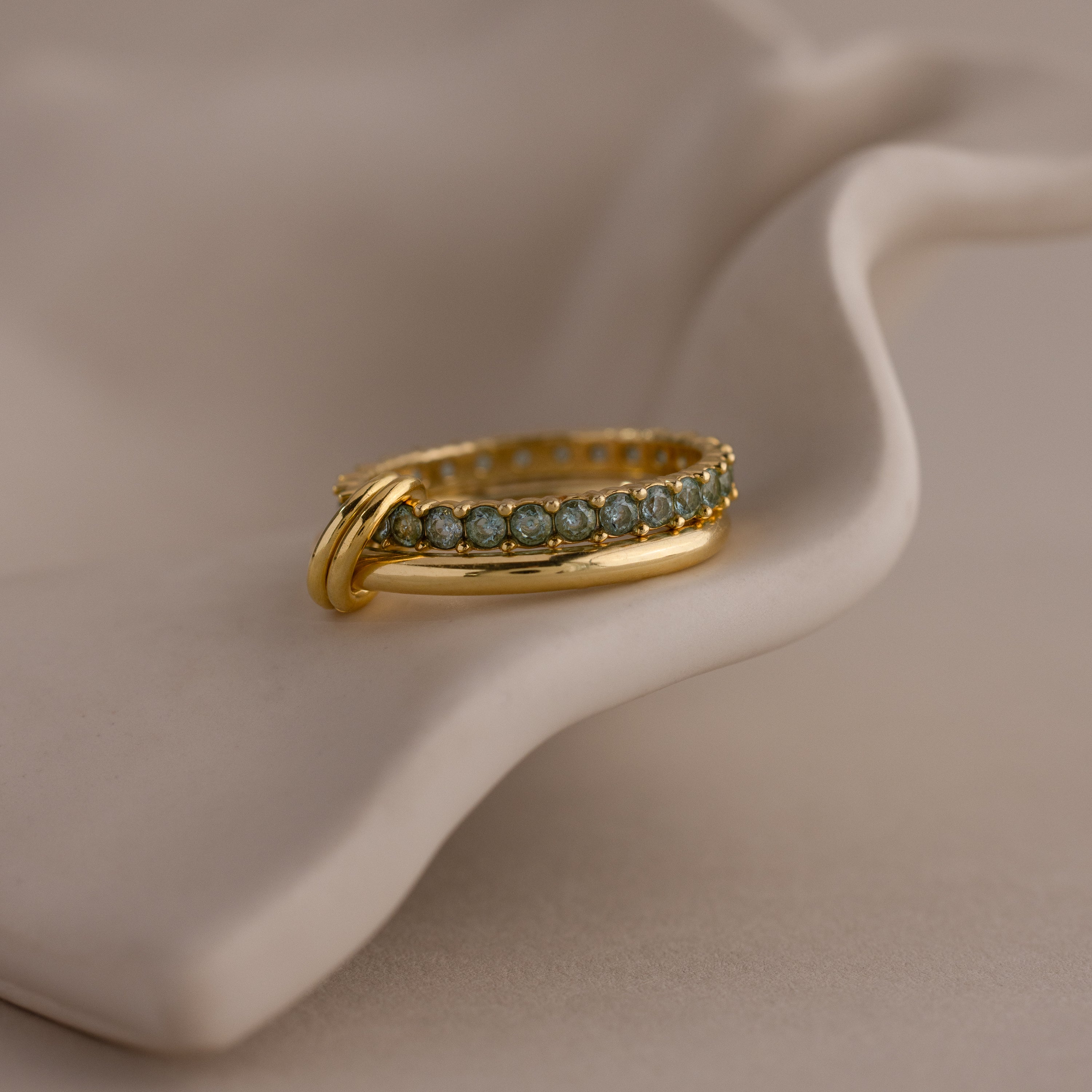 The Duo Birthstone Tennis Linked Ring Set features a row of blue-green gemstones, elegantly displayed on draped, light-colored fabric.