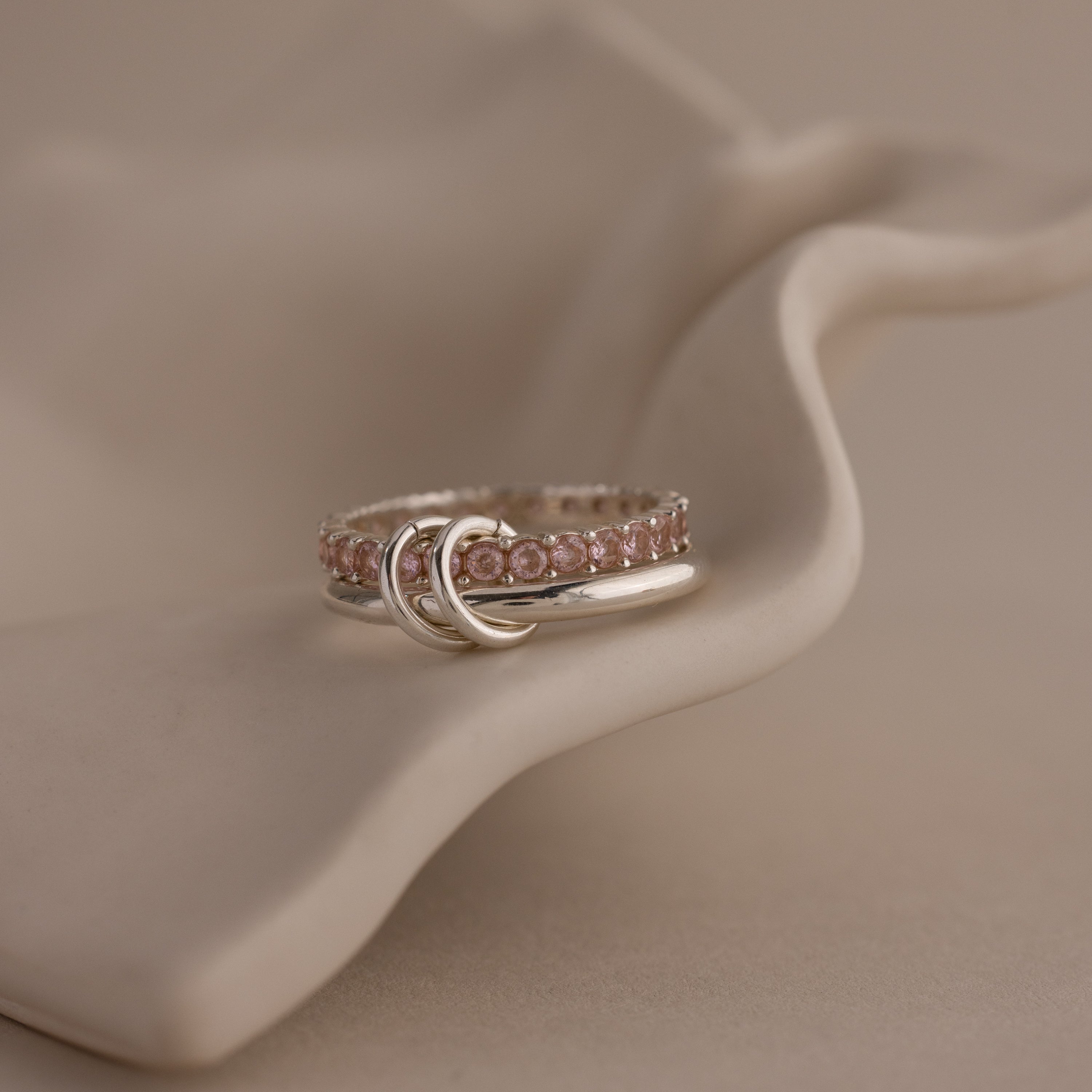 The Duo Birthstone Tennis Linked Ring Set, featuring pink gemstones and crescent moon details, rests on a wavy cream surface, offering celestial charm to your look.