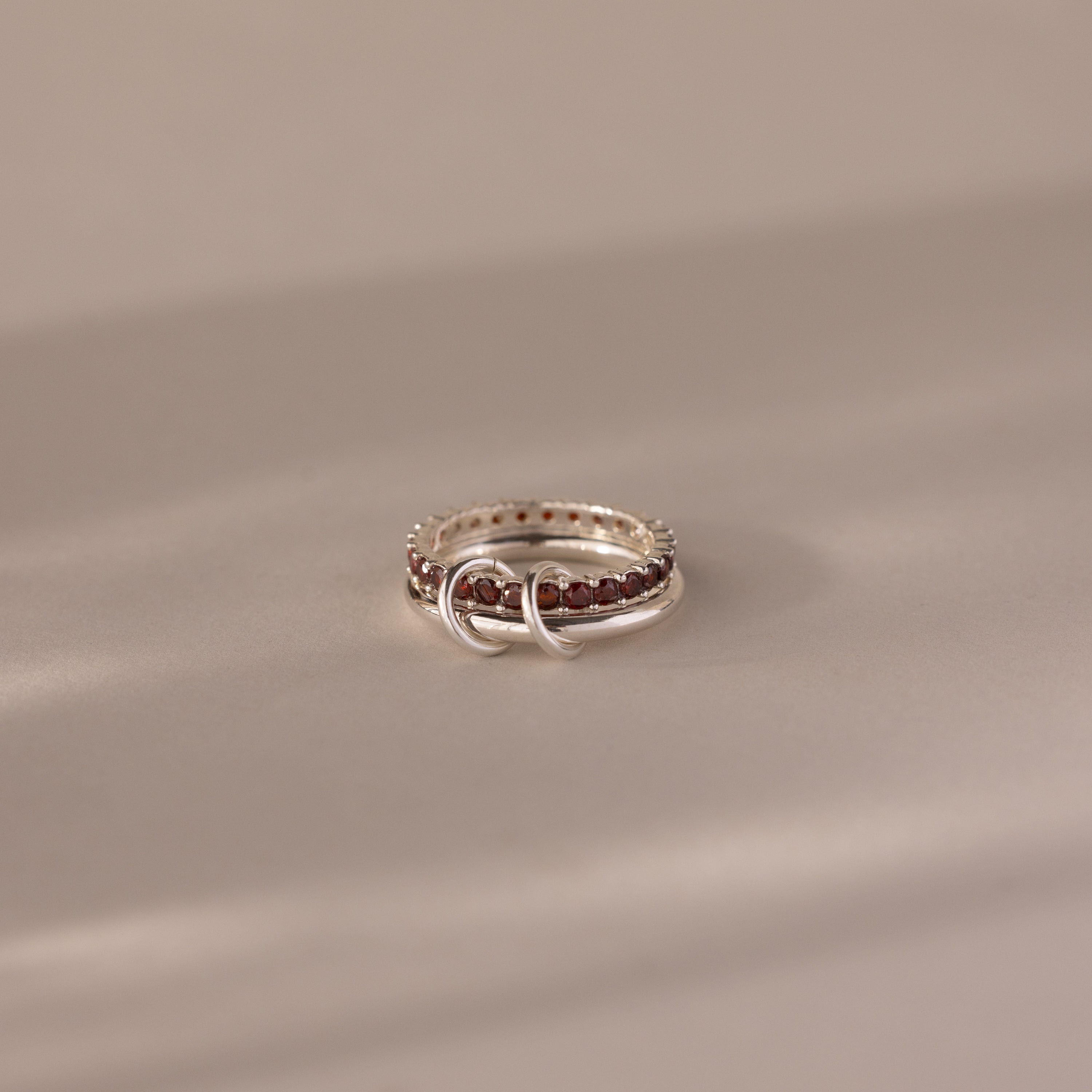 The Duo Garnet Tennis Linked Ring Set, featuring two interlocking silver loops adorned with a row of small red garnet stones, rests on a beige surface.