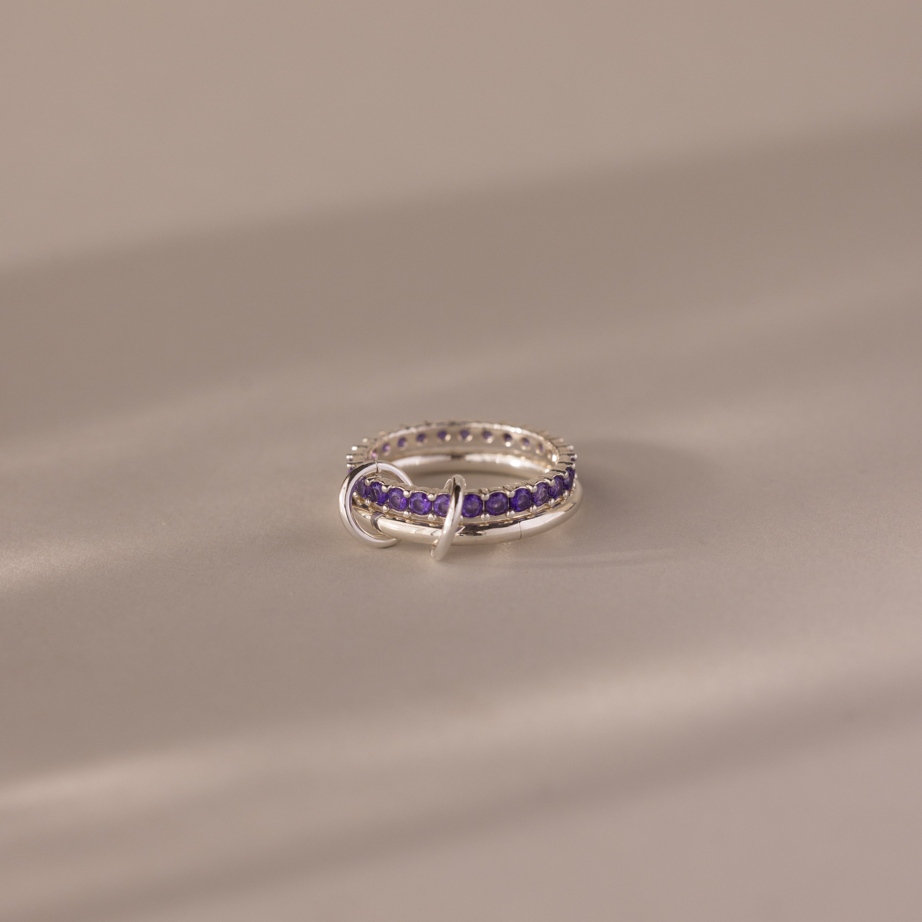 The Duo Amethyst Tennis Linked Ring Set features silver bands adorned with small amethyst gemstones in a row, shown against a soft beige background.