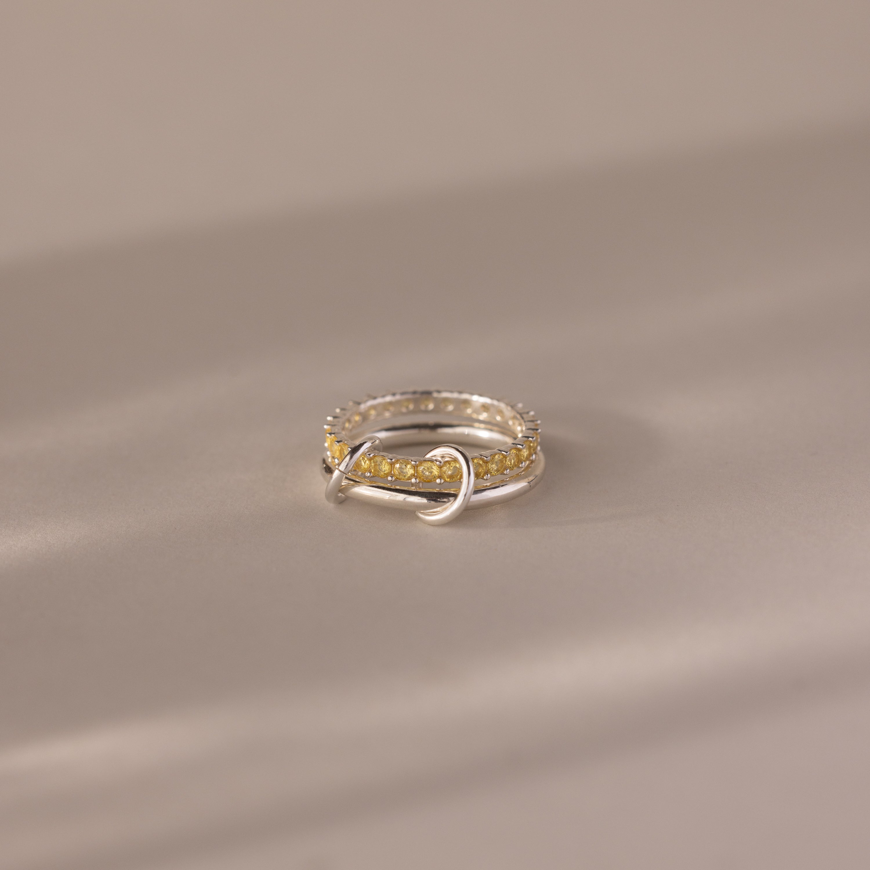 The Duo Topaz Tennis Linked Ring Set features a unique looped design with sparkling yellow gemstones, showcased on a neutral beige surface.