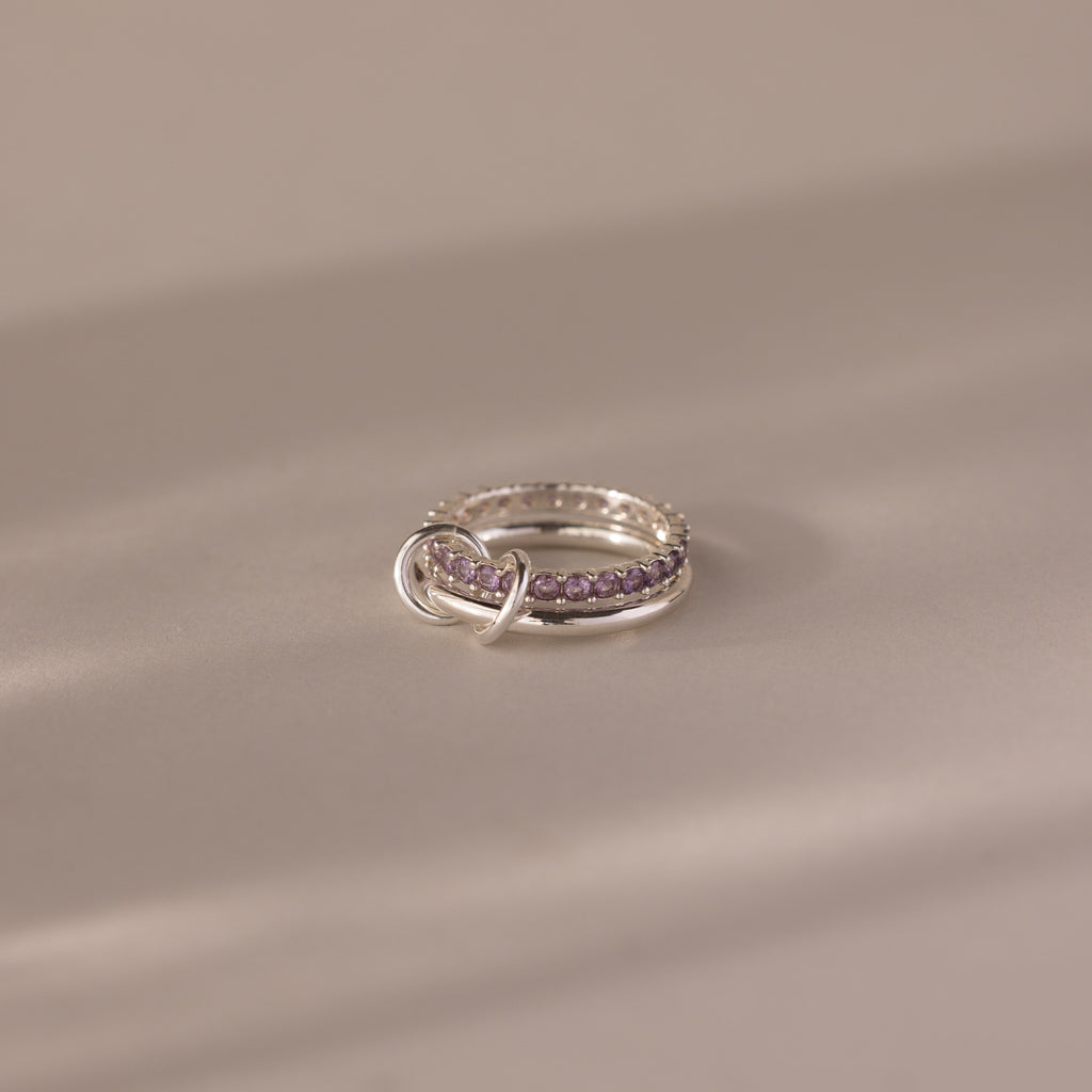 The Duo Alexandrite Tennis Linked Ring Set, featuring silver bands and purple gemstones, rests on a smooth beige surface in soft lighting.