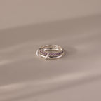The Duo Alexandrite Tennis Linked Ring Set, featuring silver bands and purple gemstones, rests on a smooth beige surface in soft lighting.
