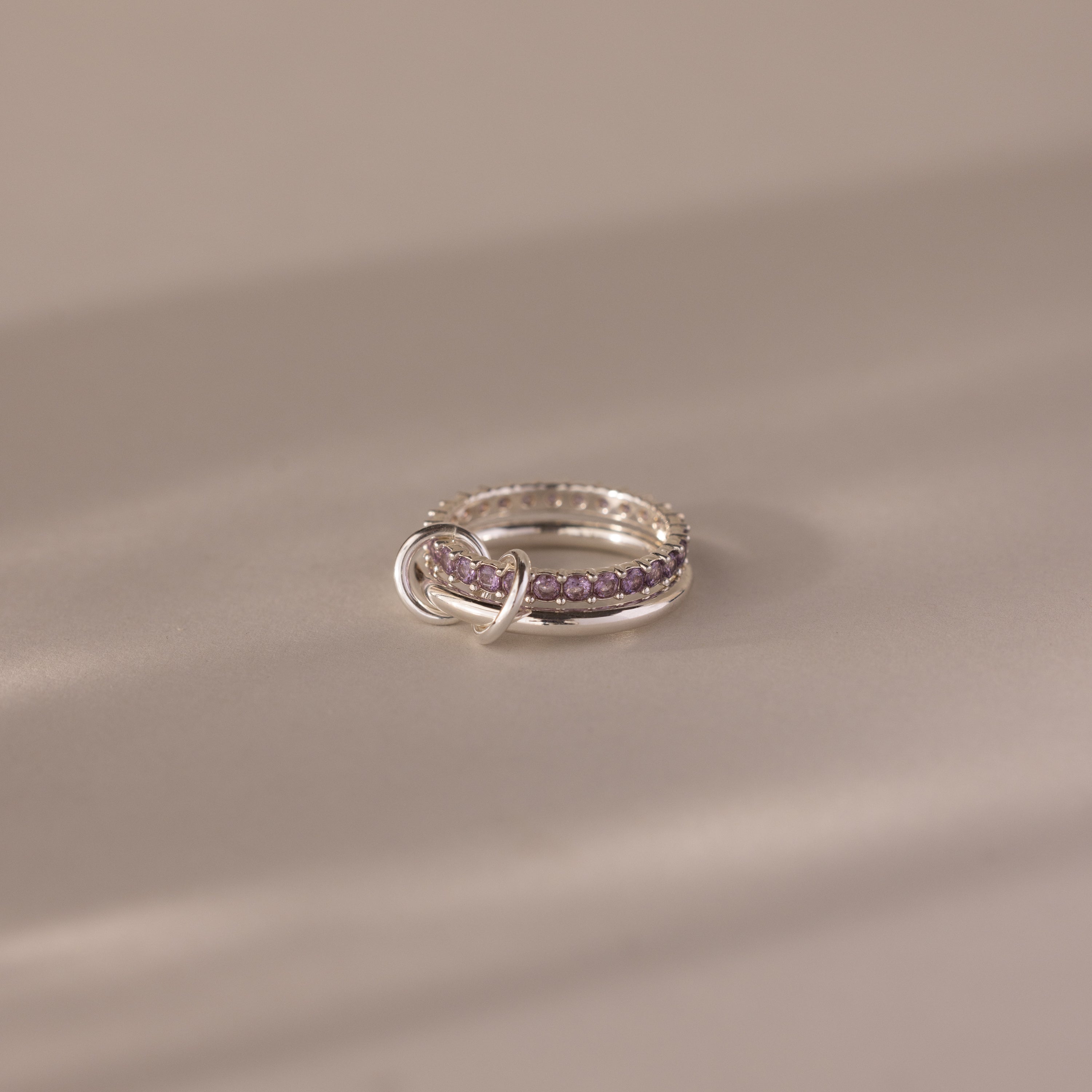 The Duo Alexandrite Tennis Linked Ring Set, featuring silver bands and purple gemstones, rests on a smooth beige surface in soft lighting.