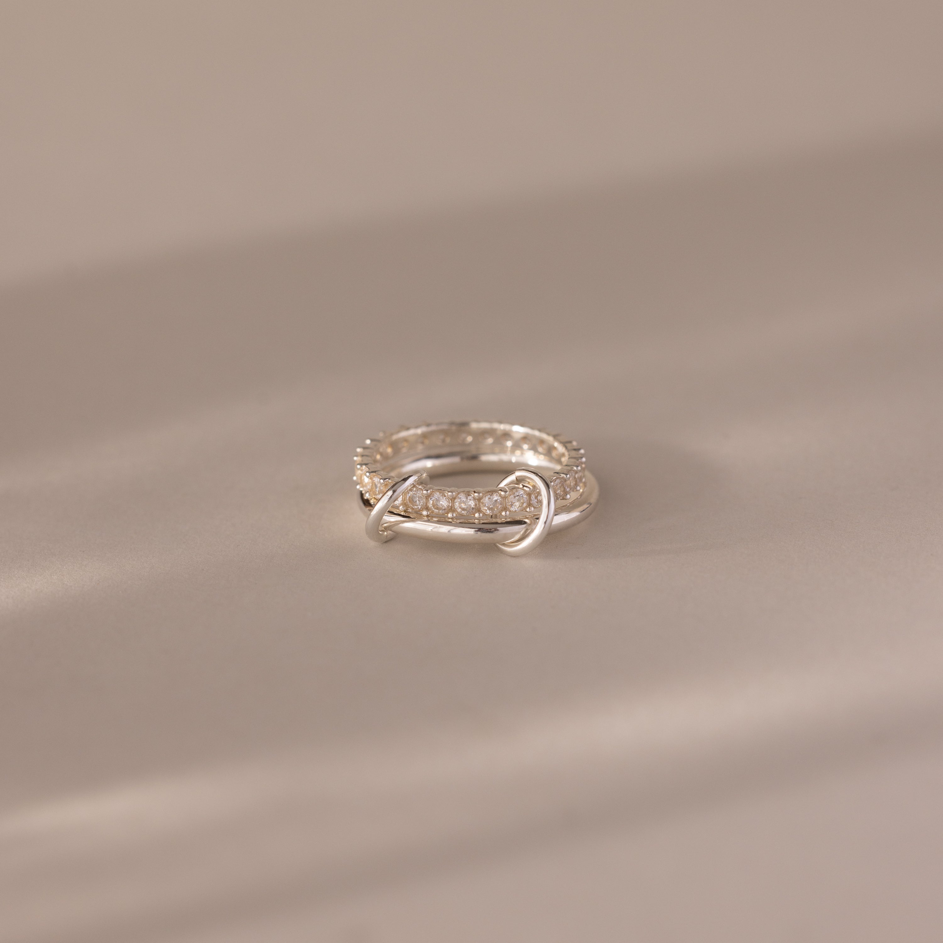 The Duo Diamond Tennis Linked Ring Set rests on a soft, beige surface, illuminated by gentle lighting.