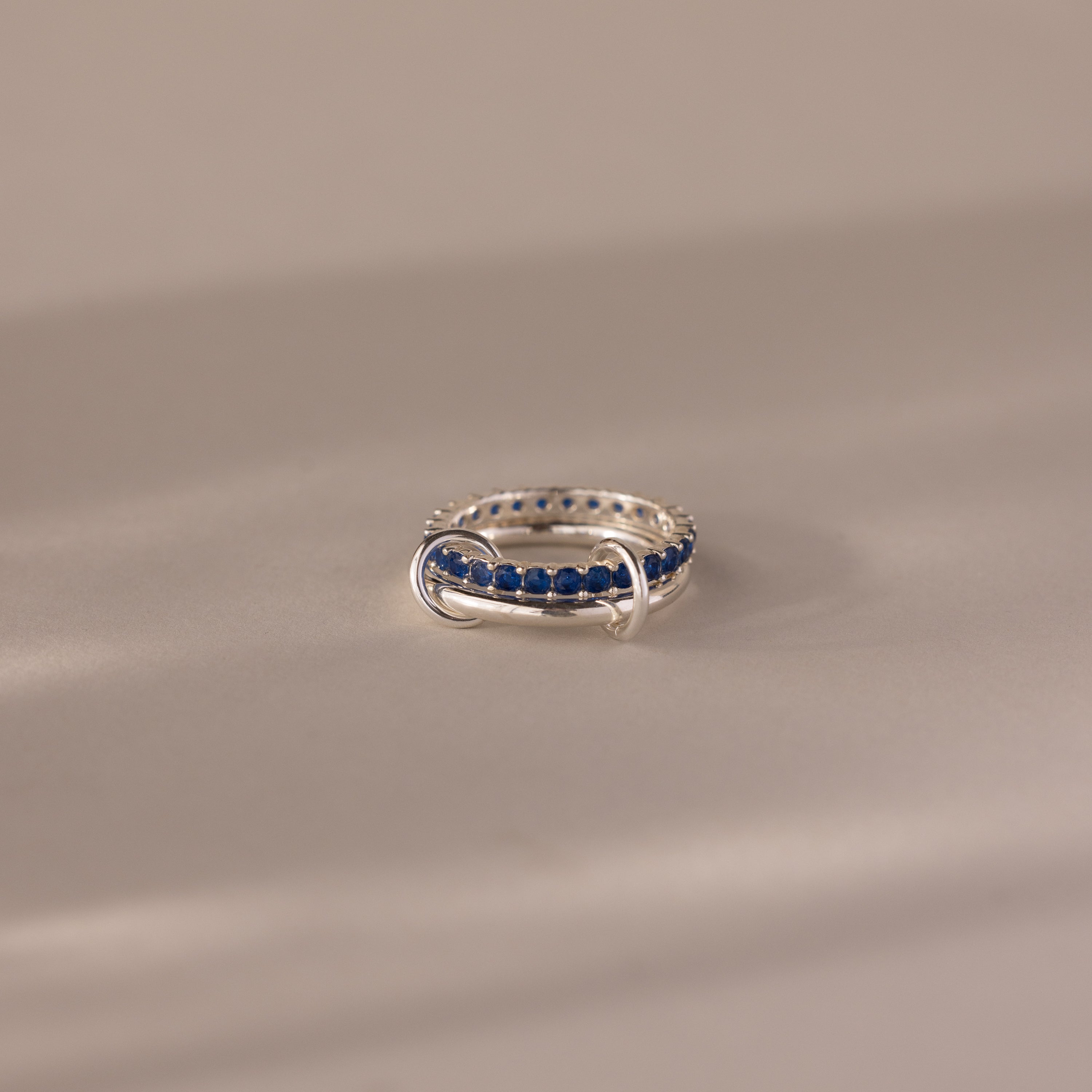The Duo Sapphire Tennis Linked Ring Set, featuring a row of blue stones in silver, is displayed on a neutral, softly lit surface.