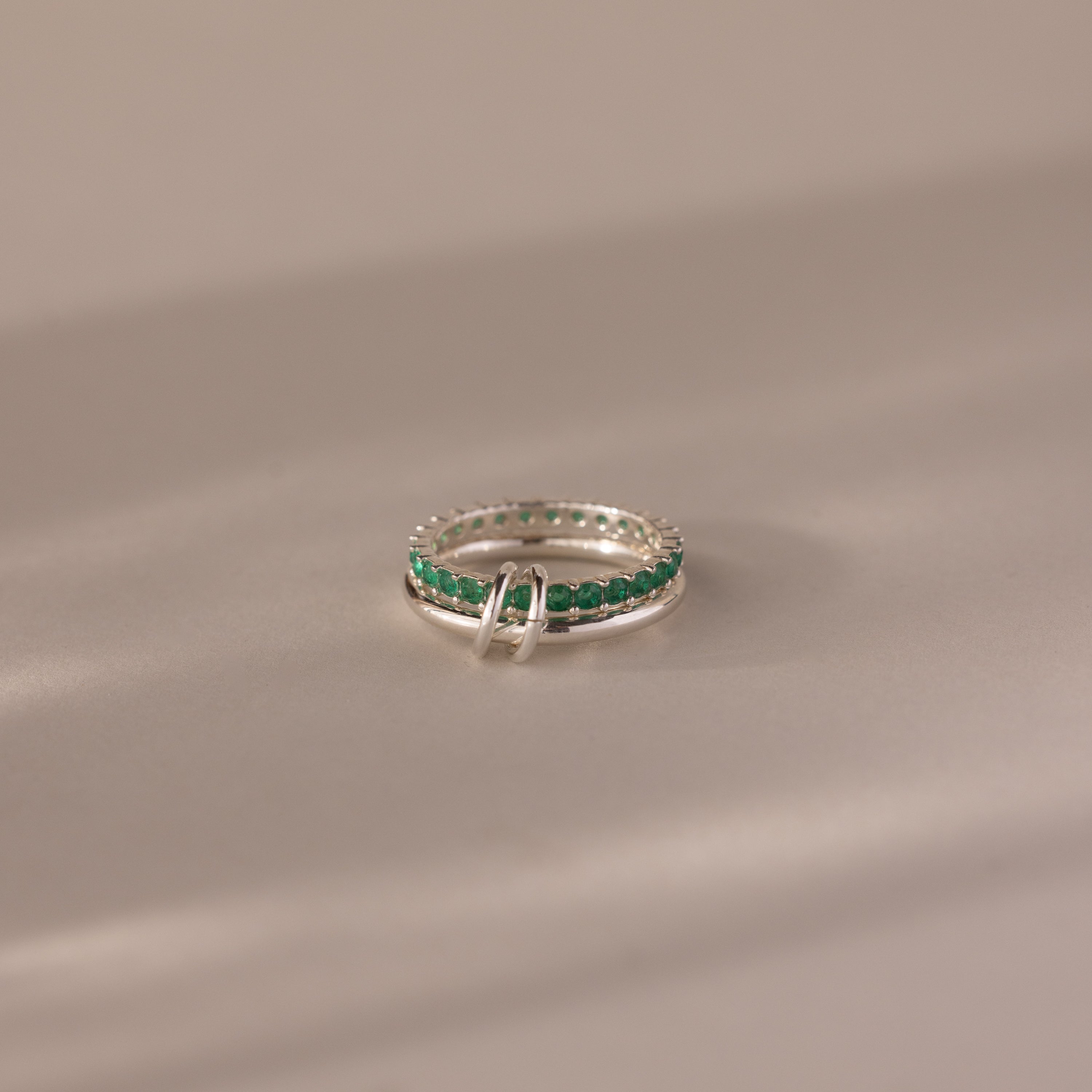 The Duo Emerald Tennis Linked Ring Set, featuring minimalist silver bands with small green stones, is displayed on a smooth, light-colored surface.