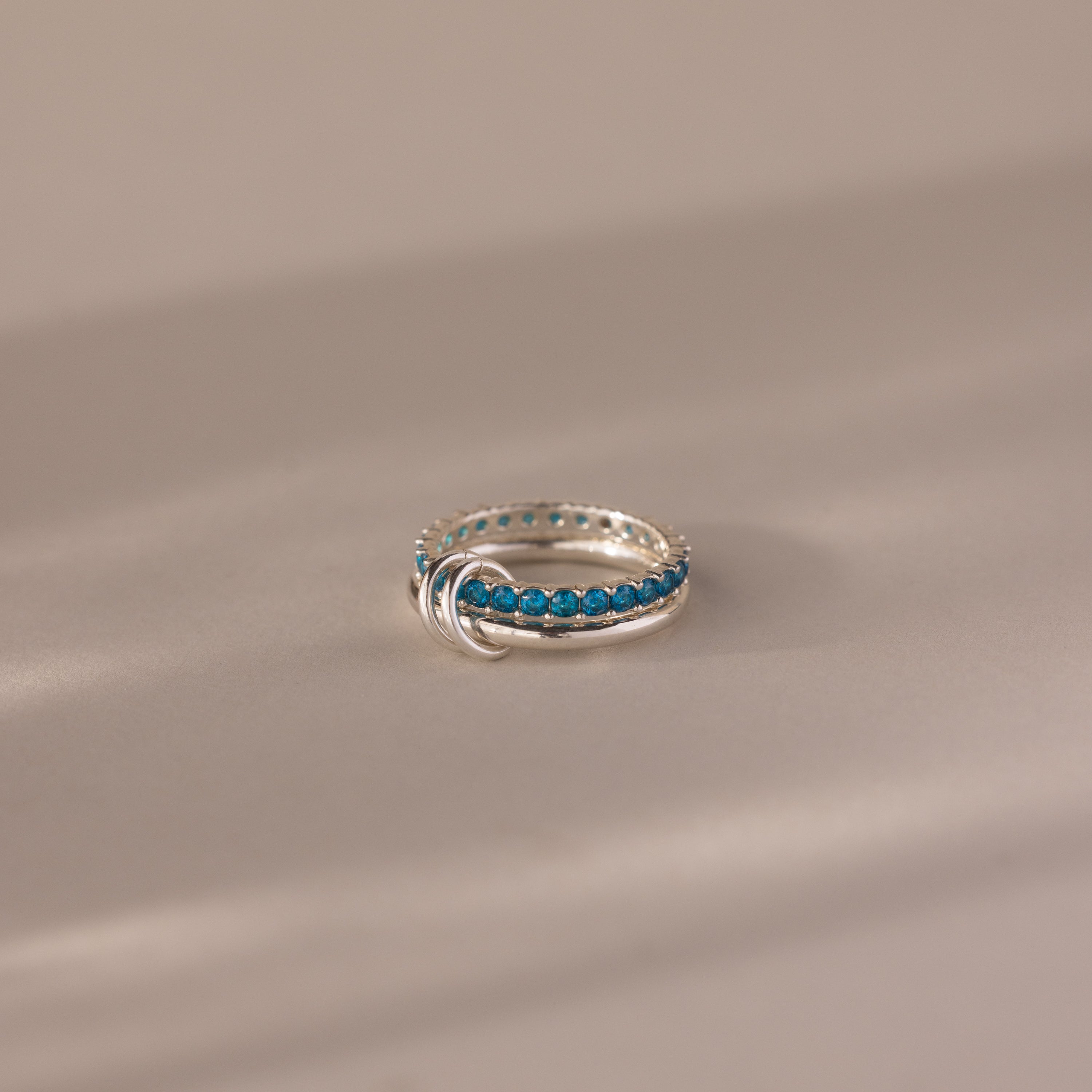 The Duo Blue Zircon Tennis Linked Ring Set, featuring a row of small blue zircon stones, is elegantly displayed on a neutral beige surface.