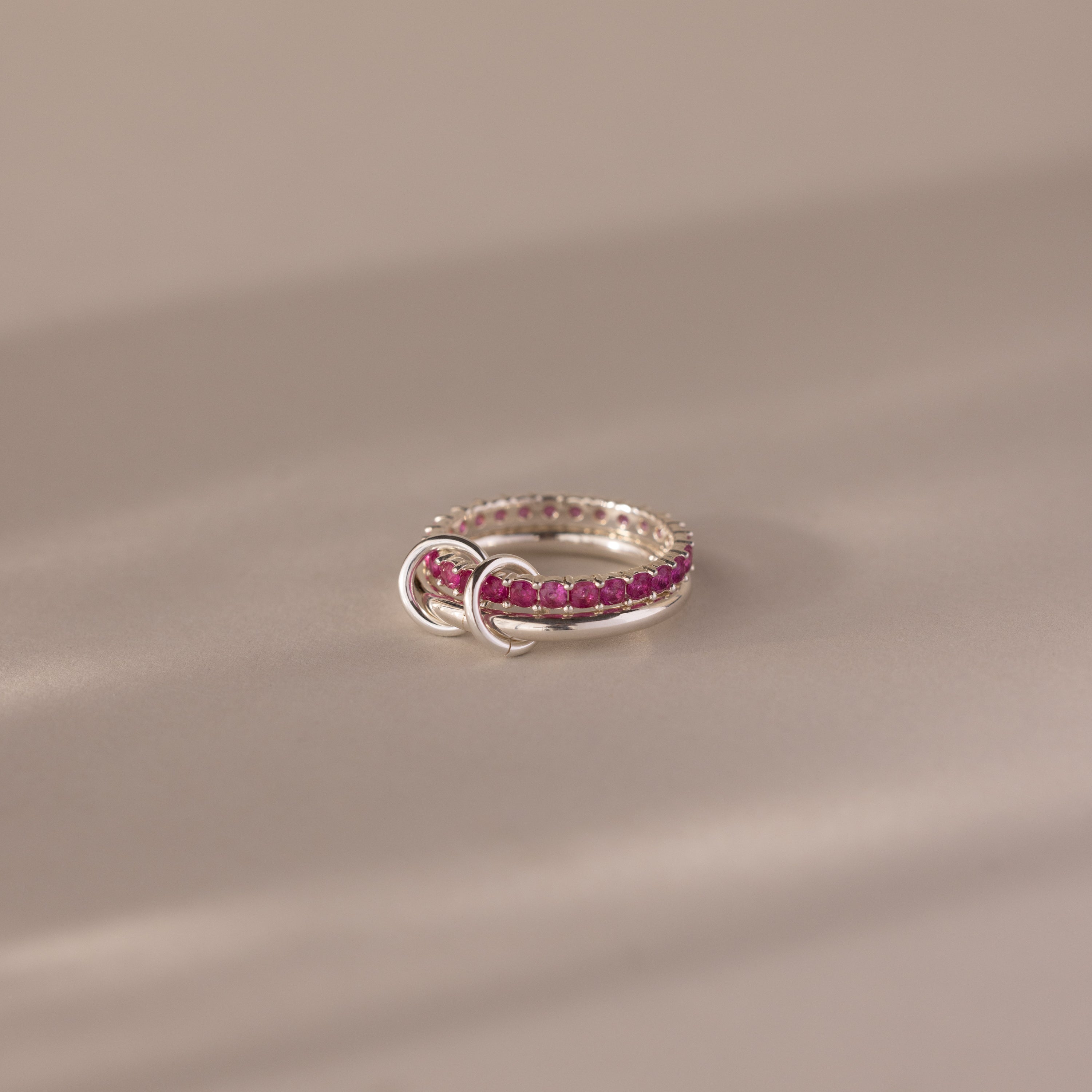 The Duo Ruby Tennis Linked Ring Set, featuring pink gemstones and two small silver loops, is displayed on a soft beige surface.